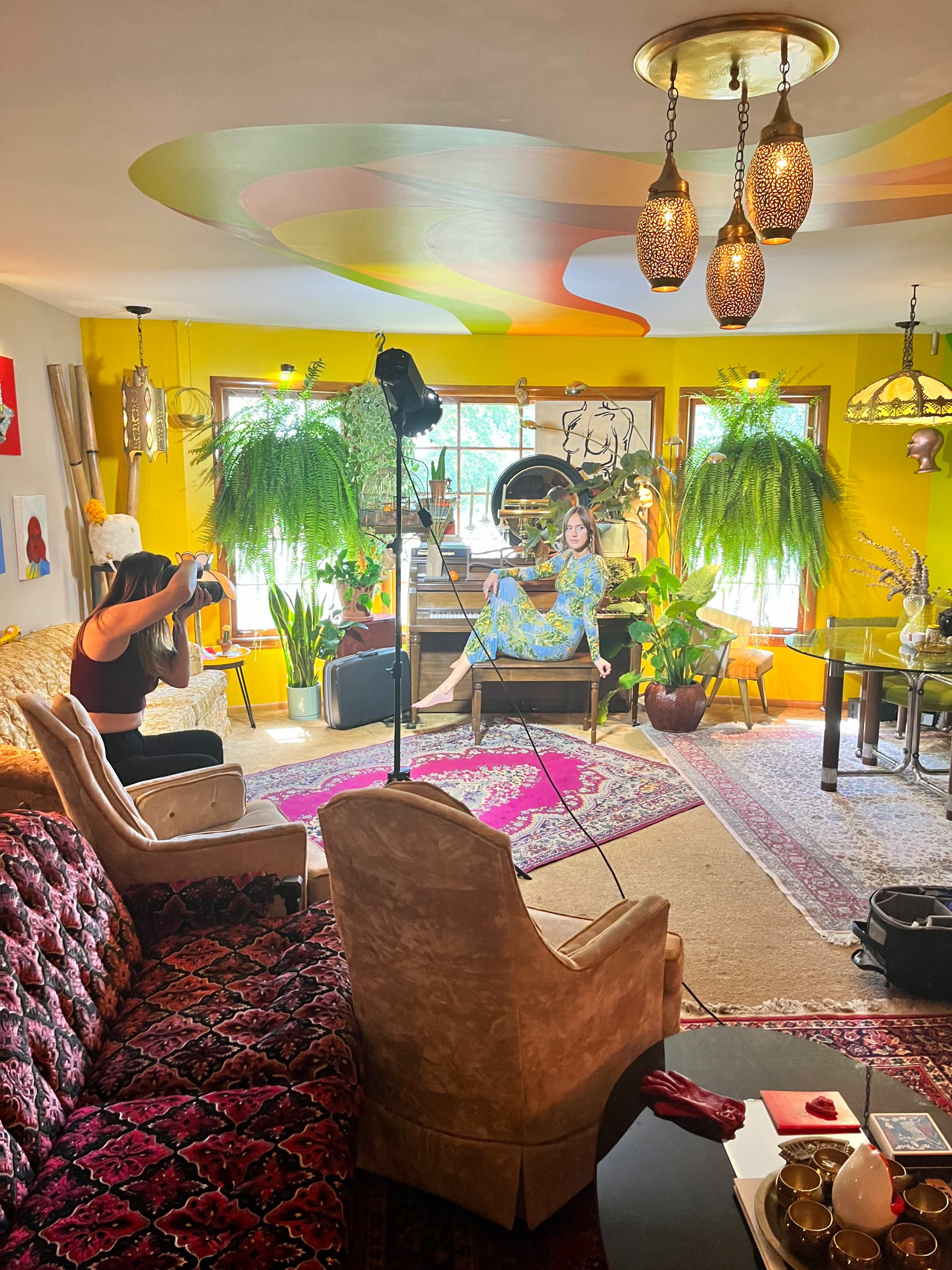 A person is photographing a model sitting in a brightly colored, plants-filled living room with eclectic decor.