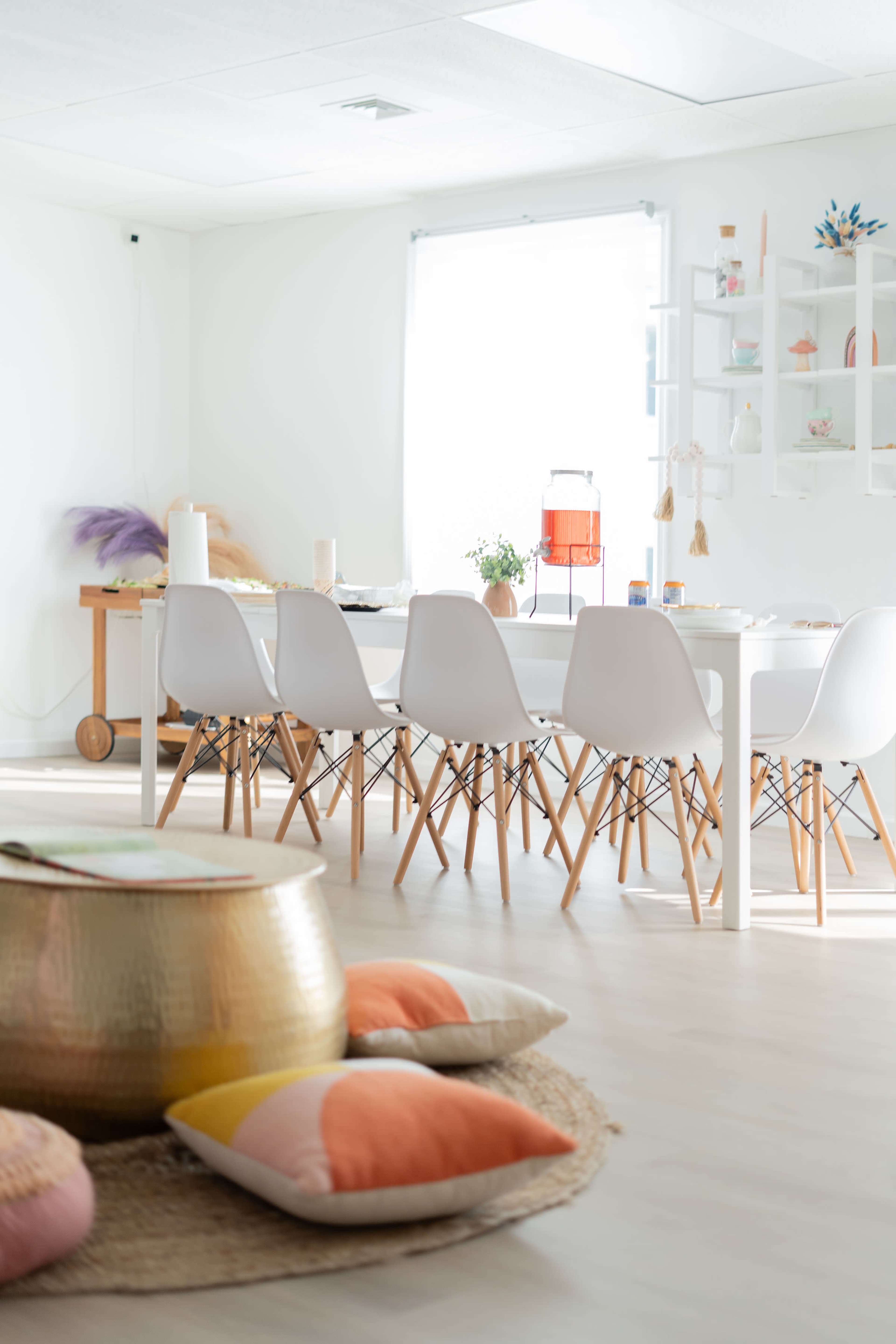 A bright, minimalist dining area features a large white table surrounded by chairs, a rug with colorful cushions, and decorative elements on the shelves.