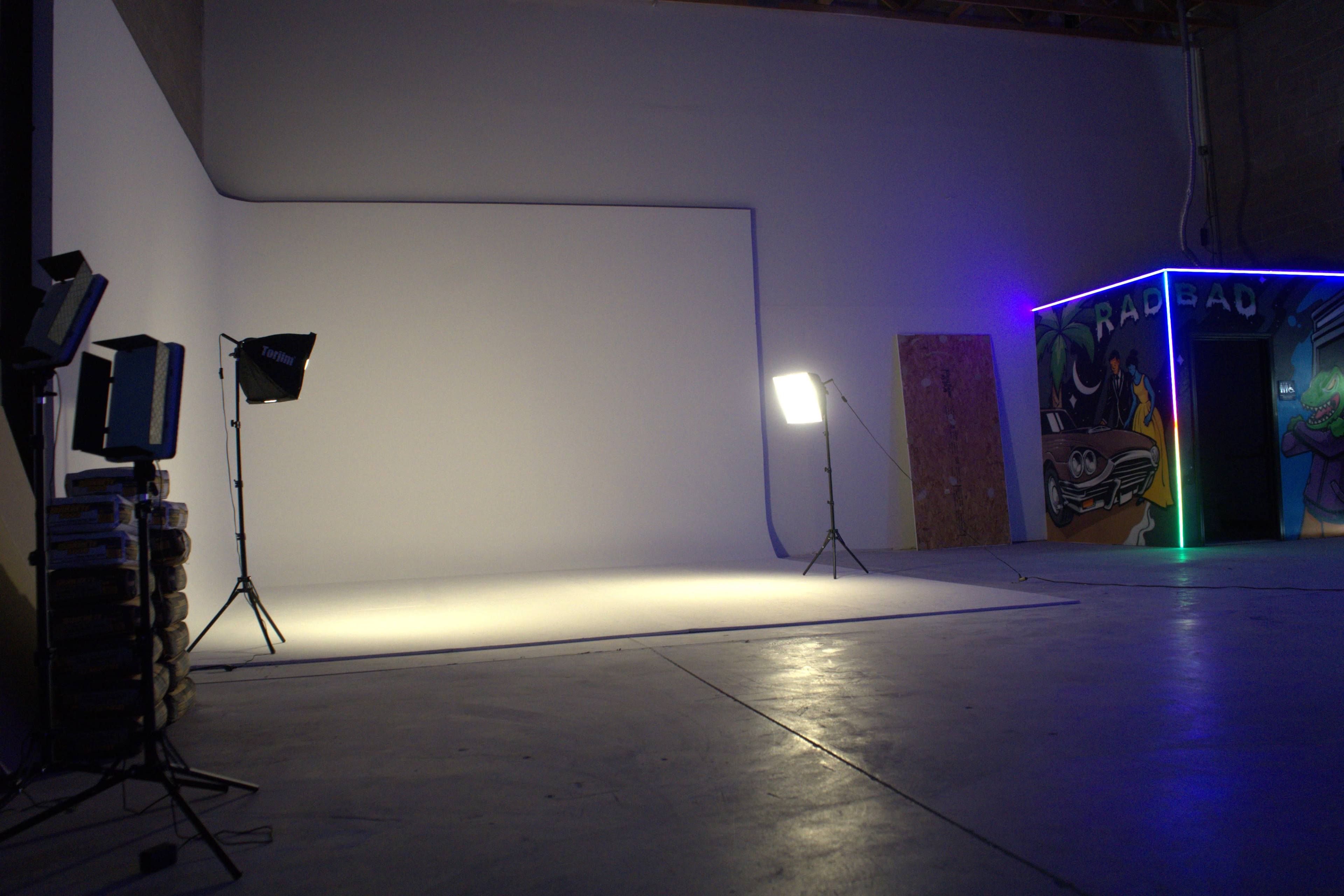 The image shows a photography studio with a blank white backdrop, lighting equipment, and a colorful mural on the side wall.