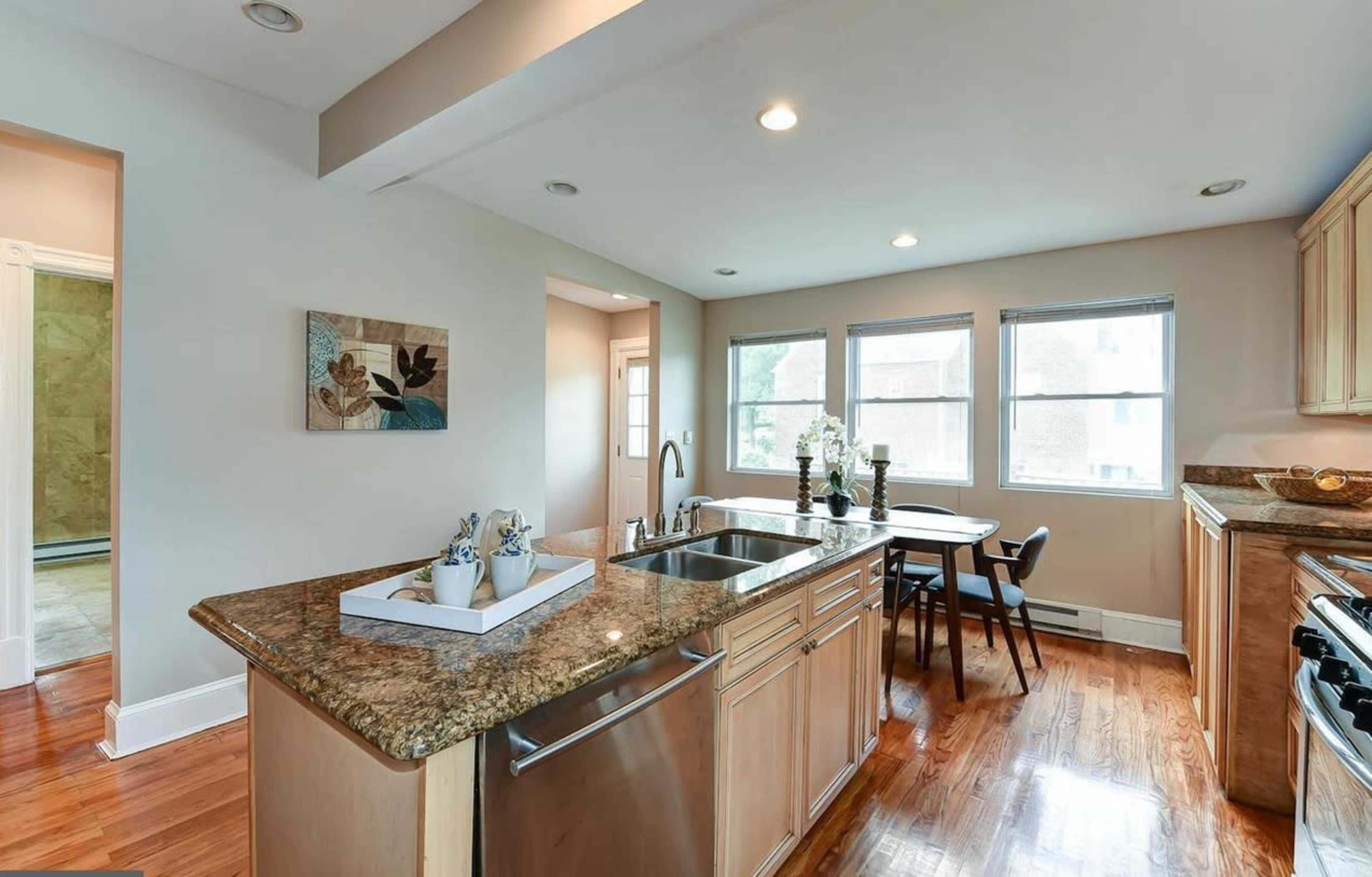A modern kitchen features a granite countertop, sink, and a dining area with a table and chairs beside large windows.