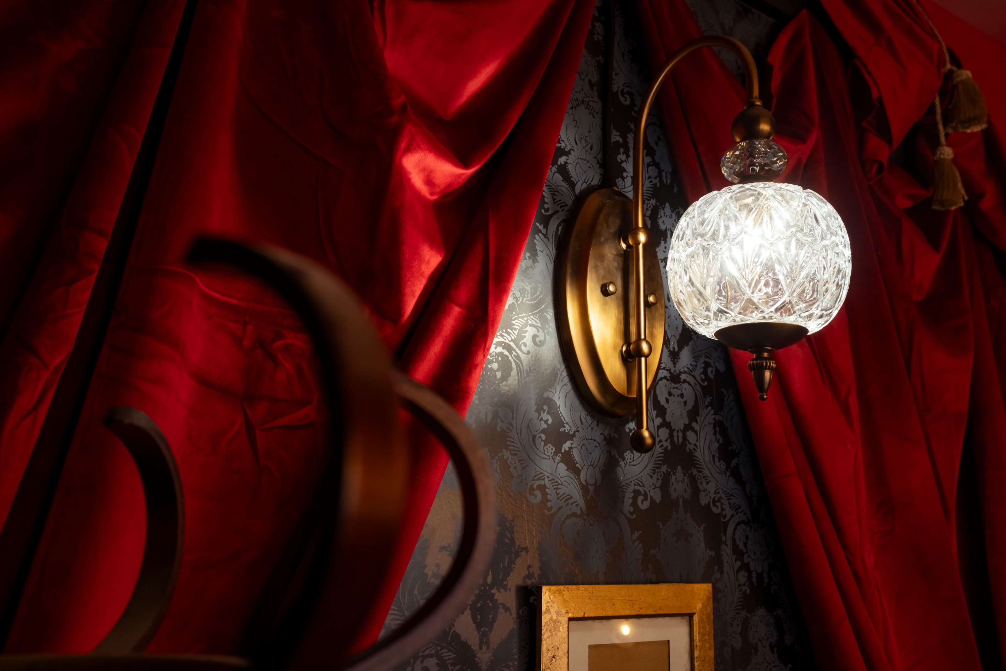 A decorative wall sconce with a crystal globe is illuminated against a backdrop of red curtains and patterned wallpaper.