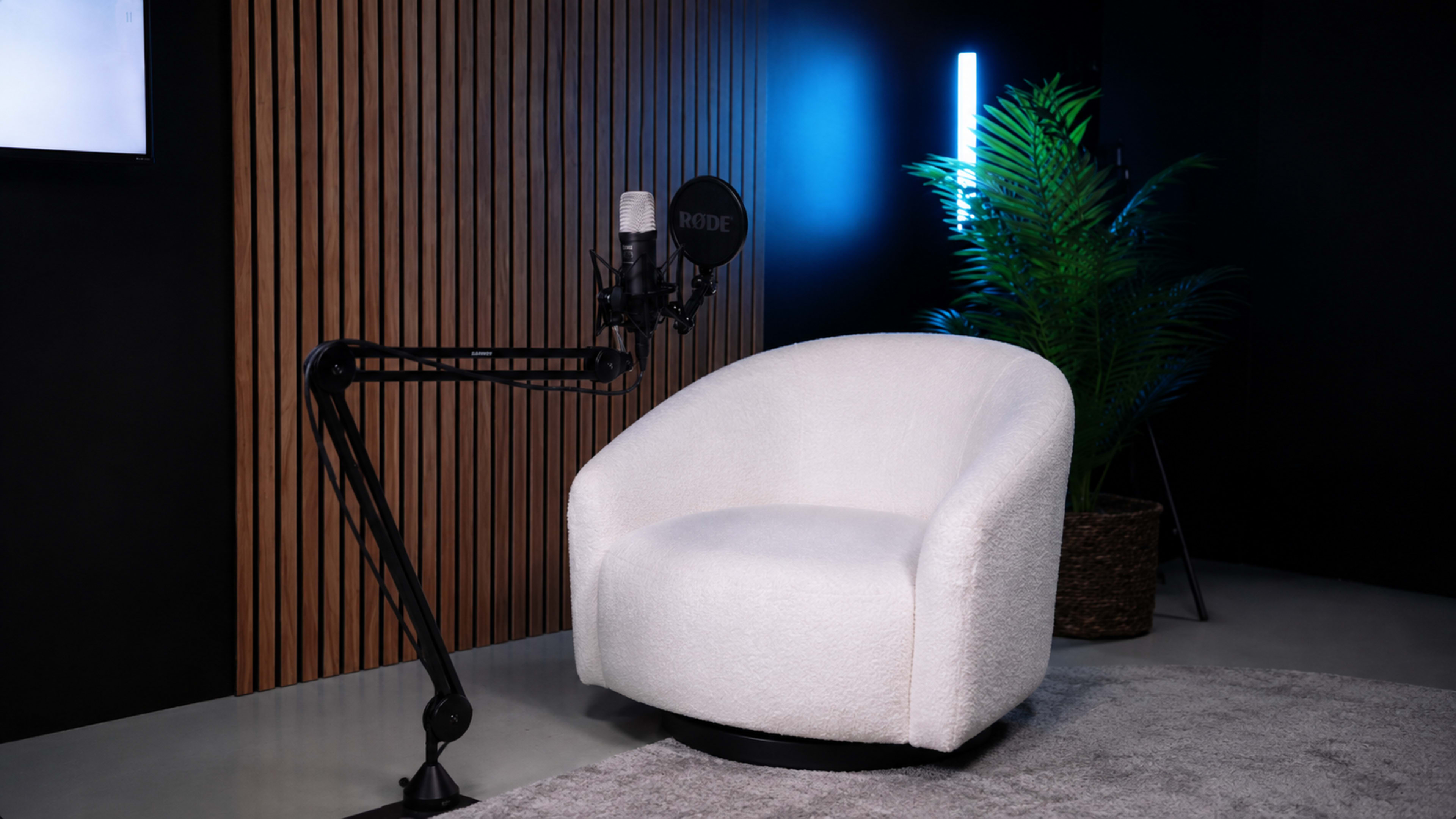 The image features a white, plush armchair positioned next to a black microphone mounted on an adjustable arm, with a leafy plant and wooden wall paneling in the background.