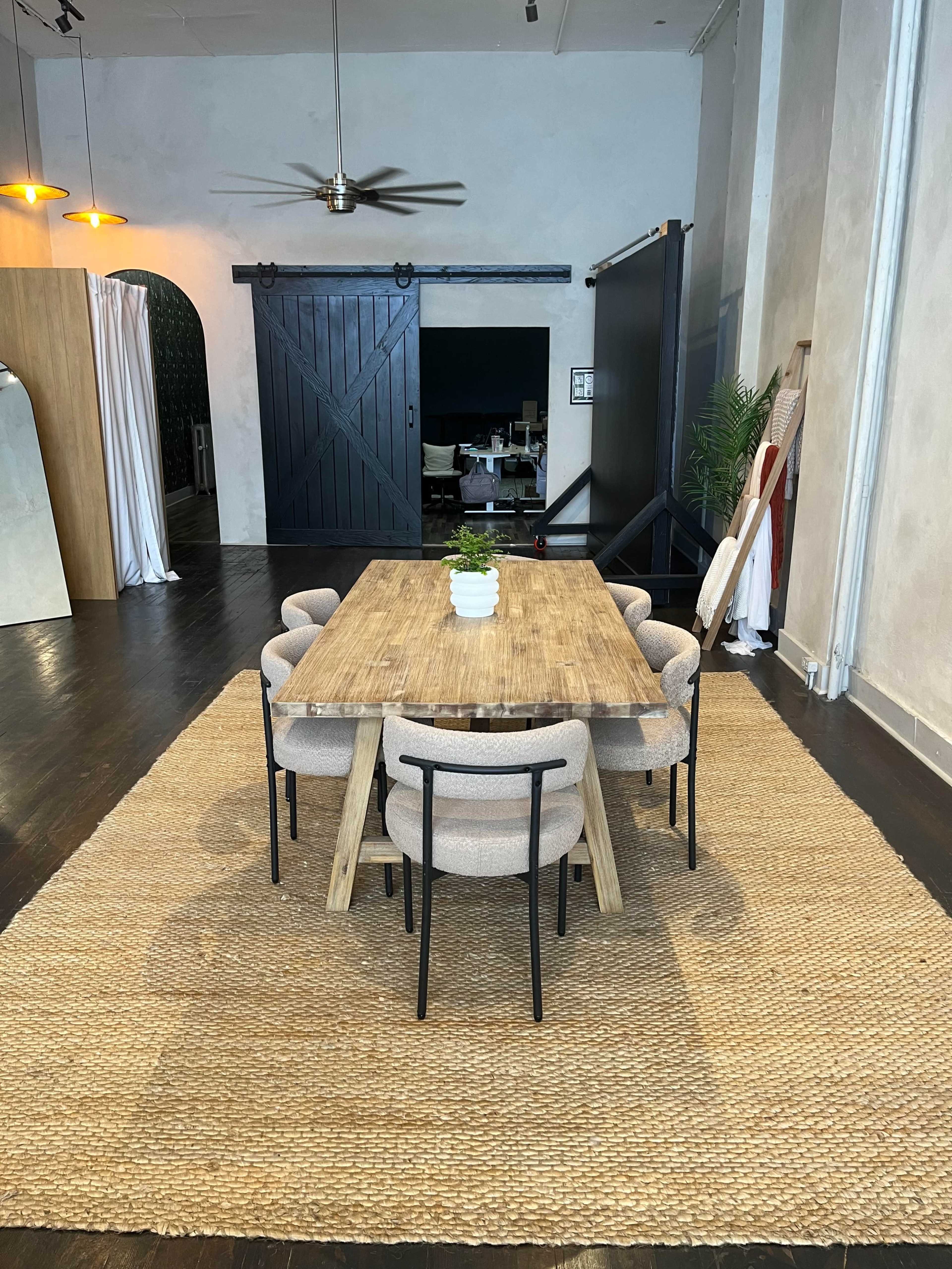 A wooden dining table surrounded by six upholstered chairs sits on a woven rug in a spacious room with a fan and dark wooden accents.