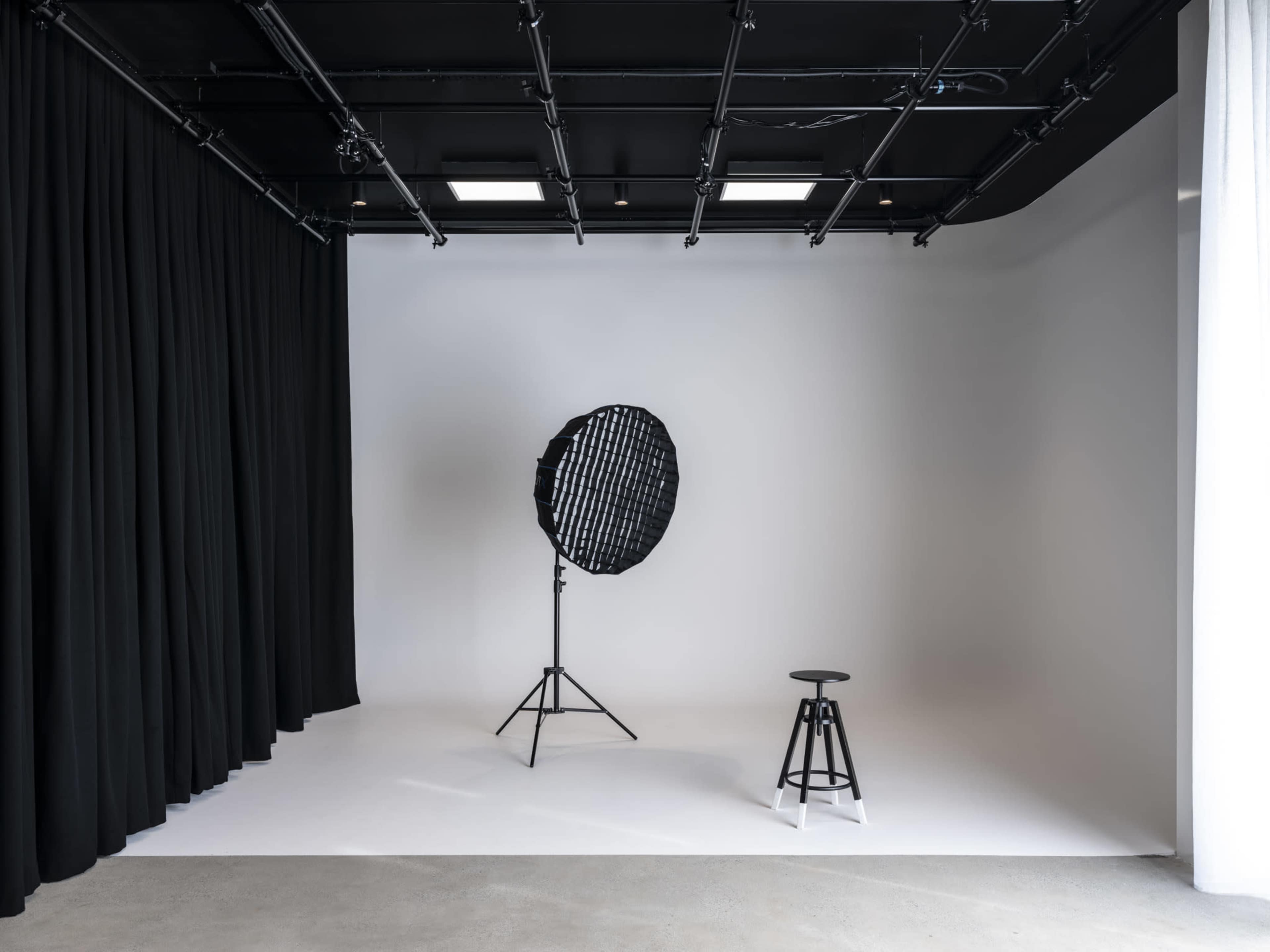 The image shows a photography studio with a large softbox light on a stand and a tall stool against a plain white backdrop.