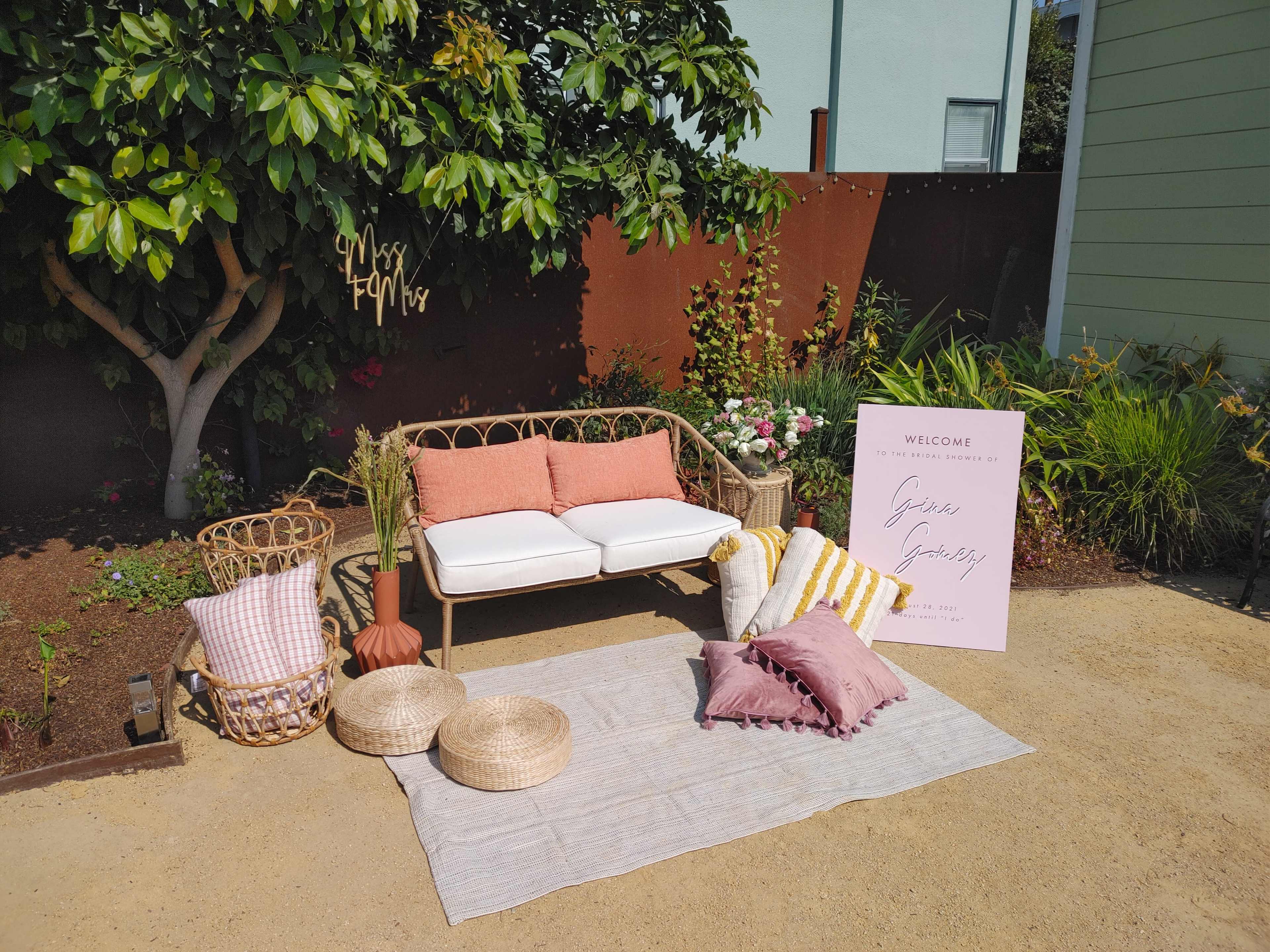 A cozy outdoor seating area features a pink and white couch, decorative pillows, woven baskets, and a welcome sign amid vibrant greenery.