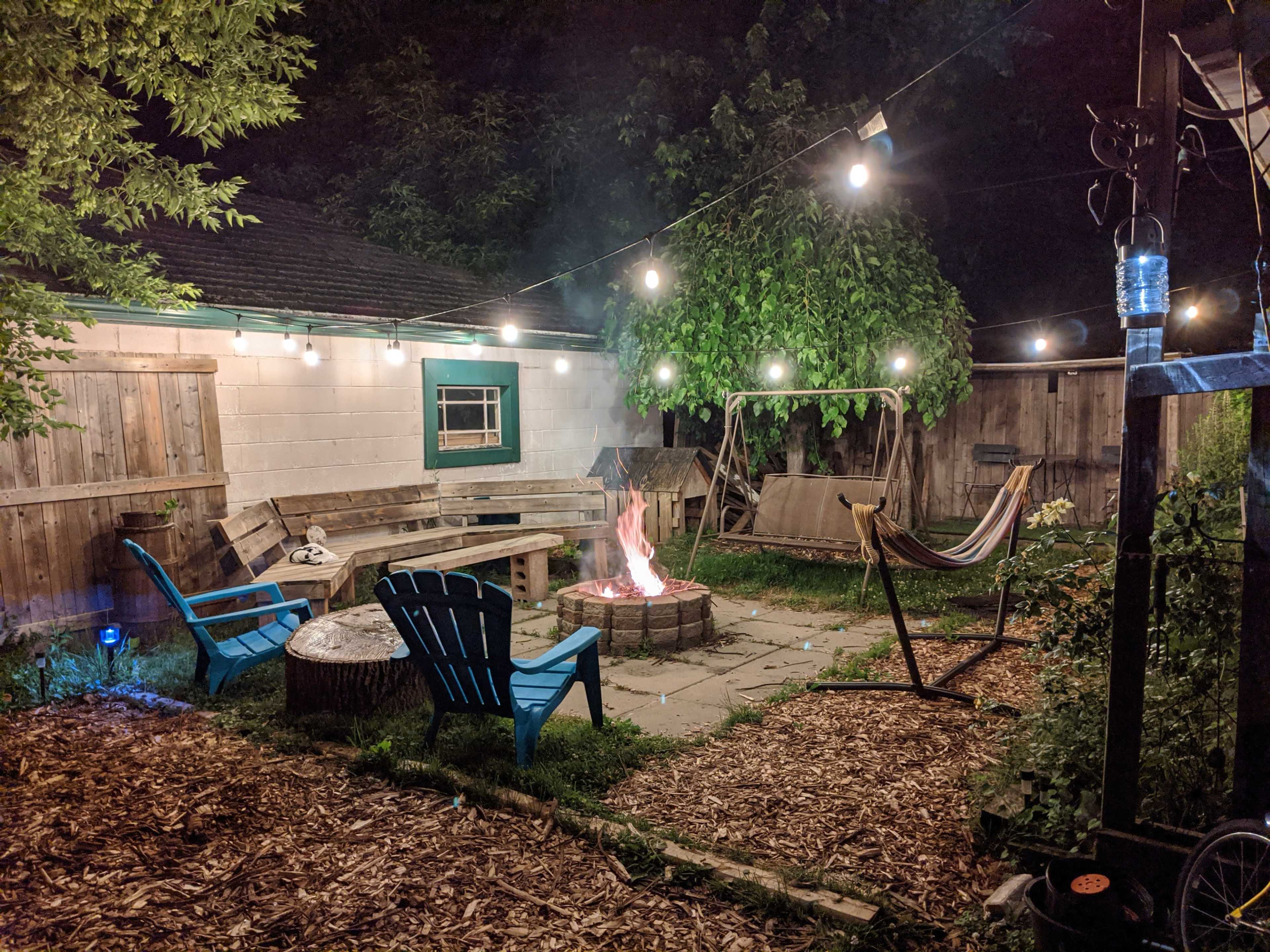 A backyard scene features a fire pit surrounded by seating, illuminated by string lights overhead, with a hammock and swing in the background.