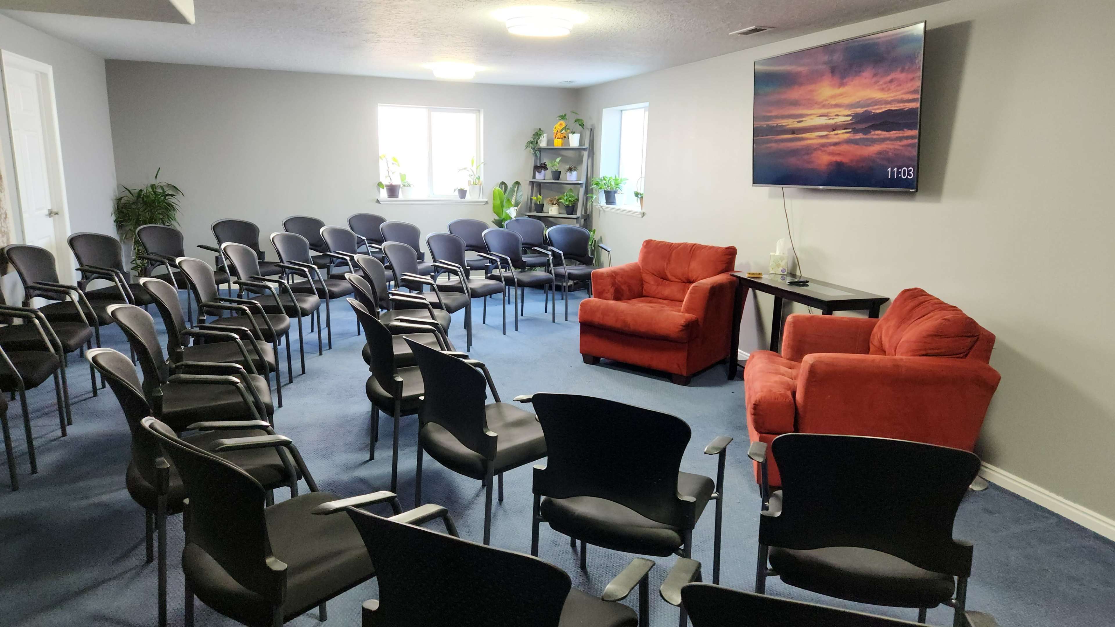 The image shows a meeting room arranged with several black chairs facing a red couch and a second chair, along with a large wall-mounted TV displaying a sunset.