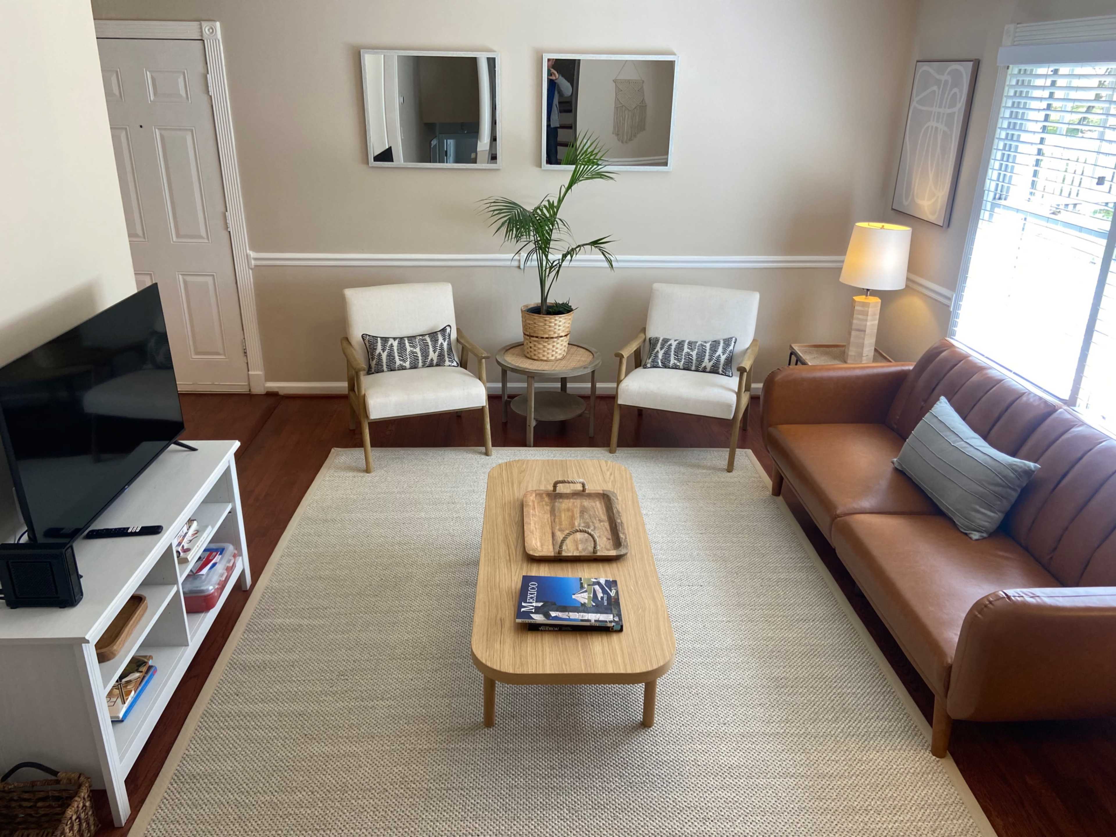 A living room features a brown sofa, two light chairs, a coffee table, a television stand, and a potted plant near a window.
