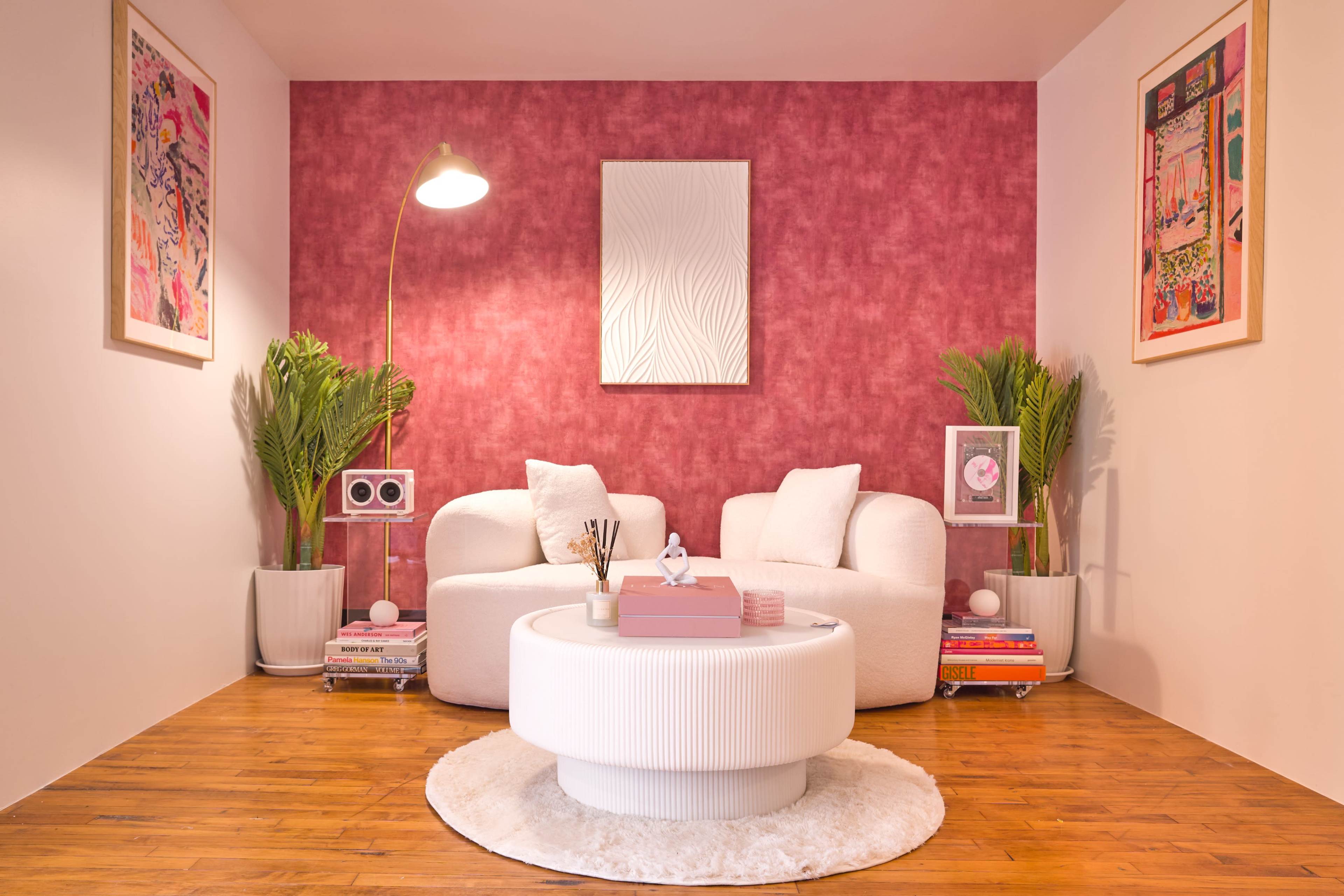 The image features a cozy living room with two white upholstered chairs, a round coffee table, and a vibrant pink textured wall, complemented by decorative plants and artwork.