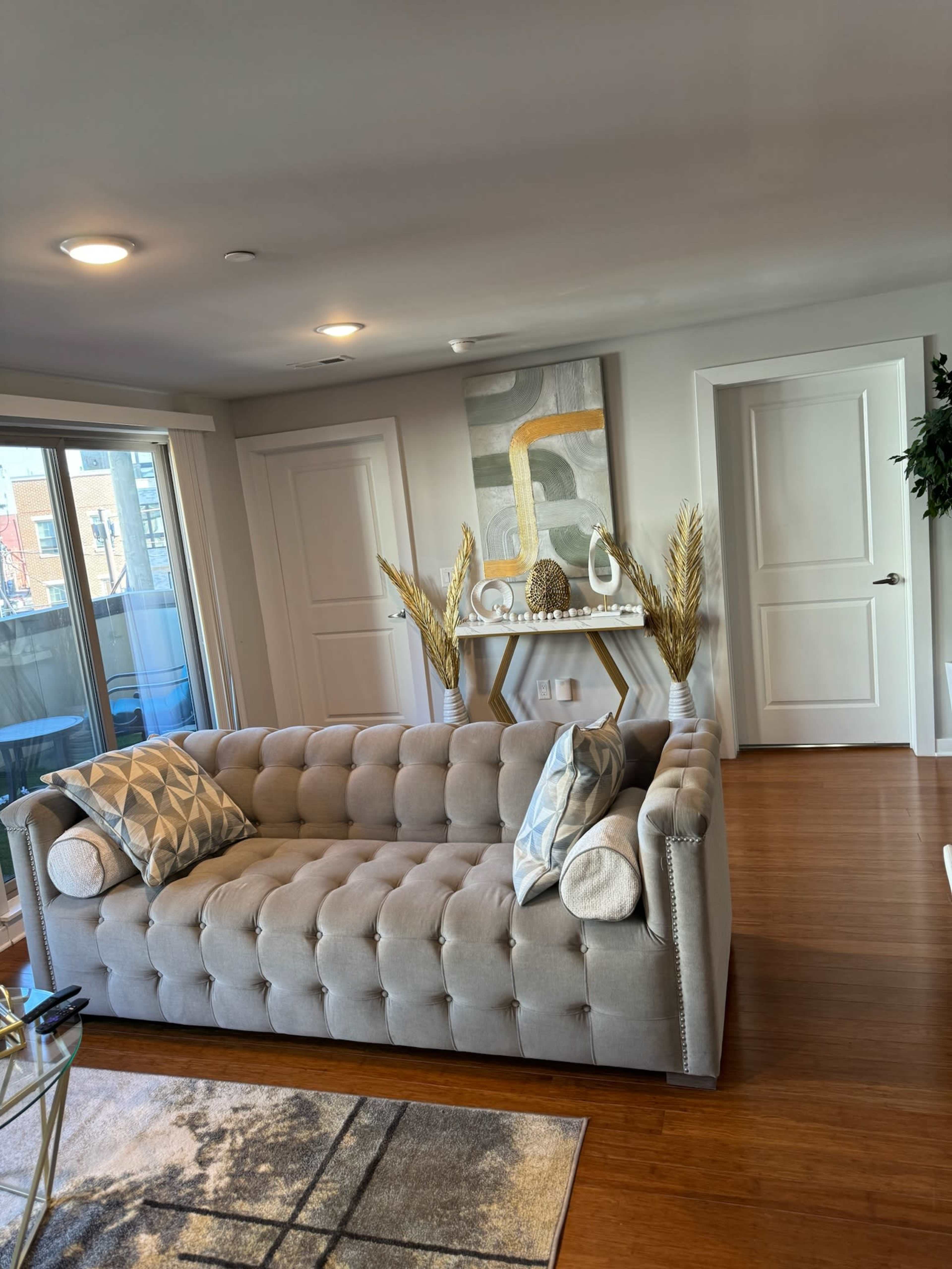 The image shows a modern living room with a light gray tufted sofa, decorative pillows, a console table with a decorative arrangement, and large windows allowing natural light.