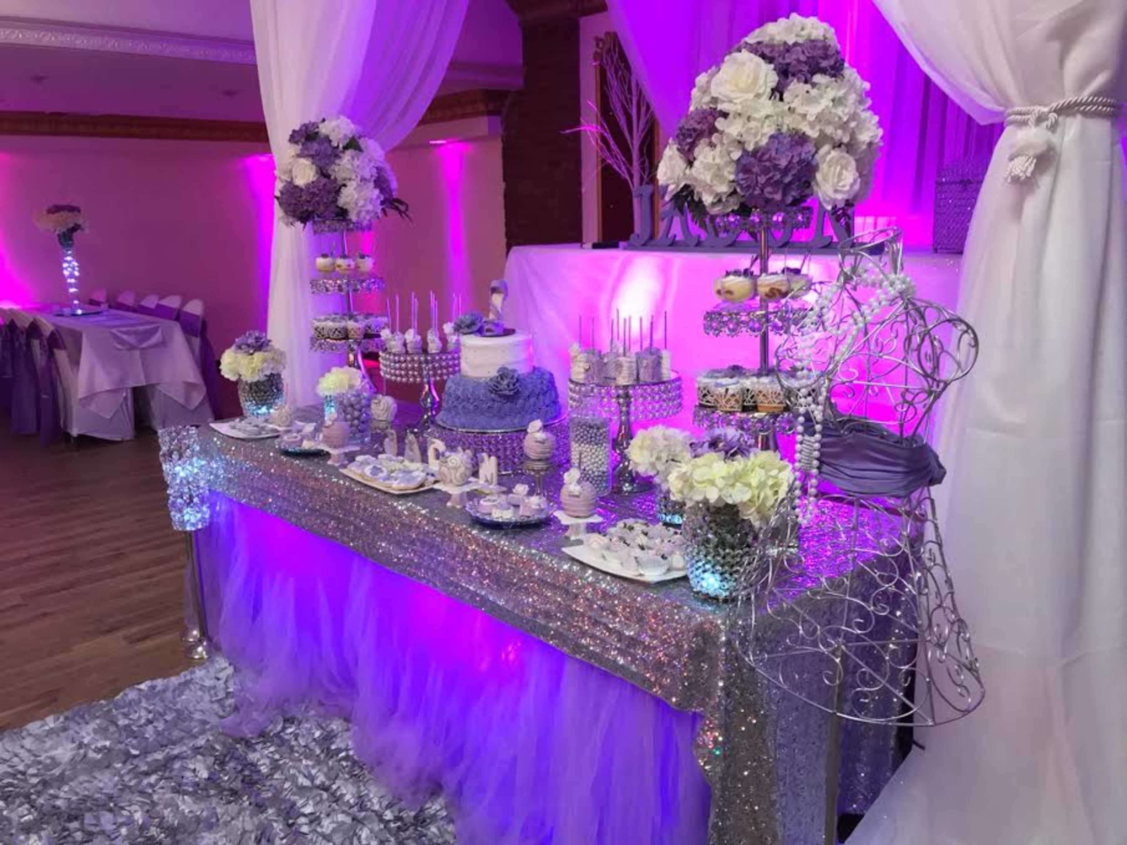 A decorated dessert table featuring a lavender cake, various sweets, and floral arrangements, set up in a venue with purple lighting.