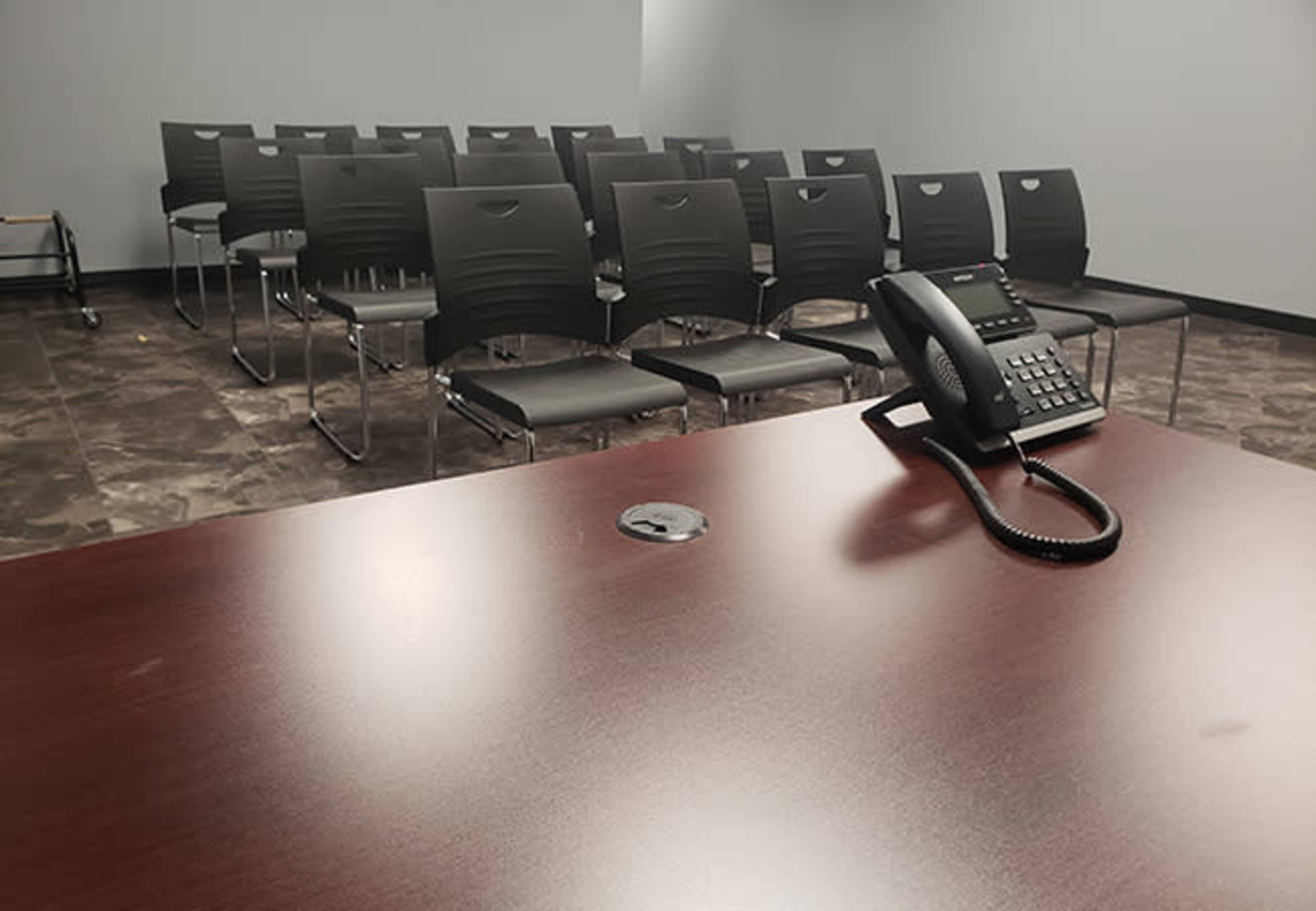 The image shows an empty room with a wooden table, a black telephone, and rows of black chairs in the background.