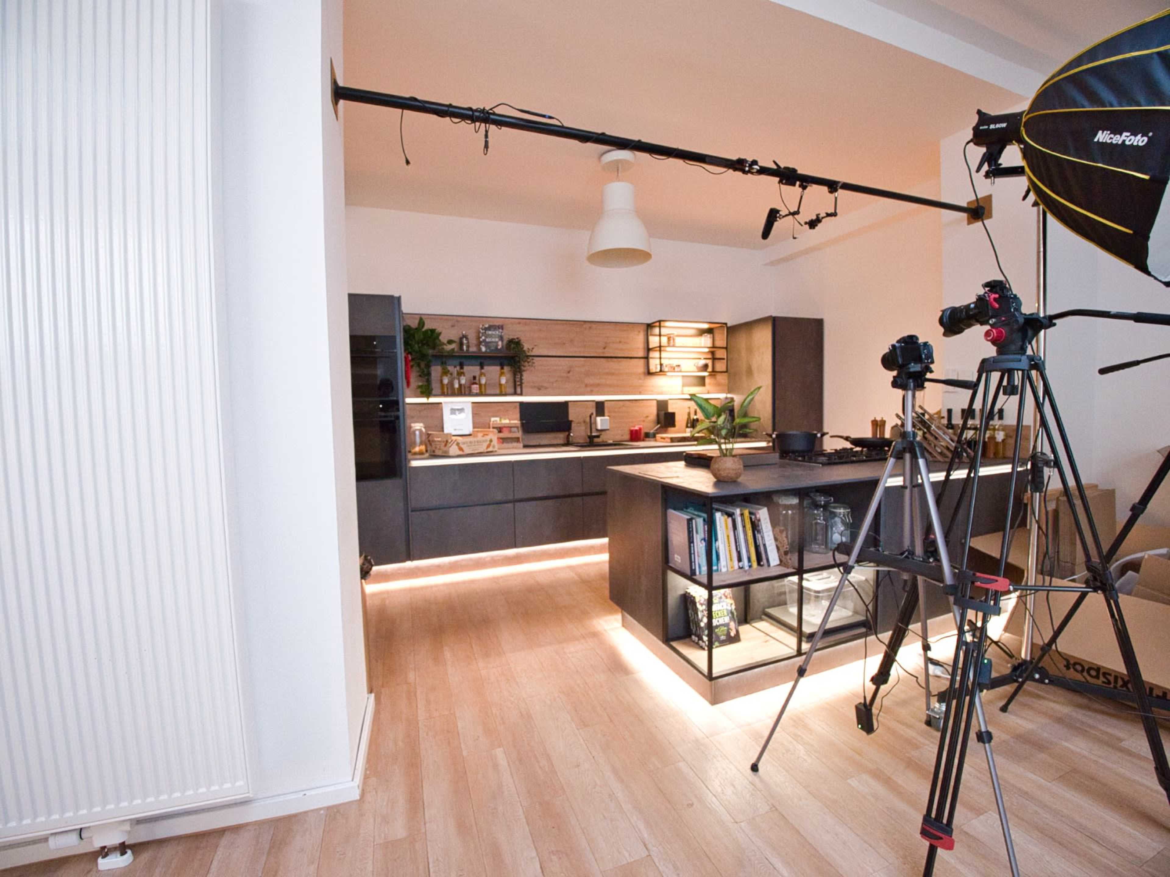 A modern kitchen is shown with dark cabinetry, a wooden accent wall, and media equipment set up for filming.