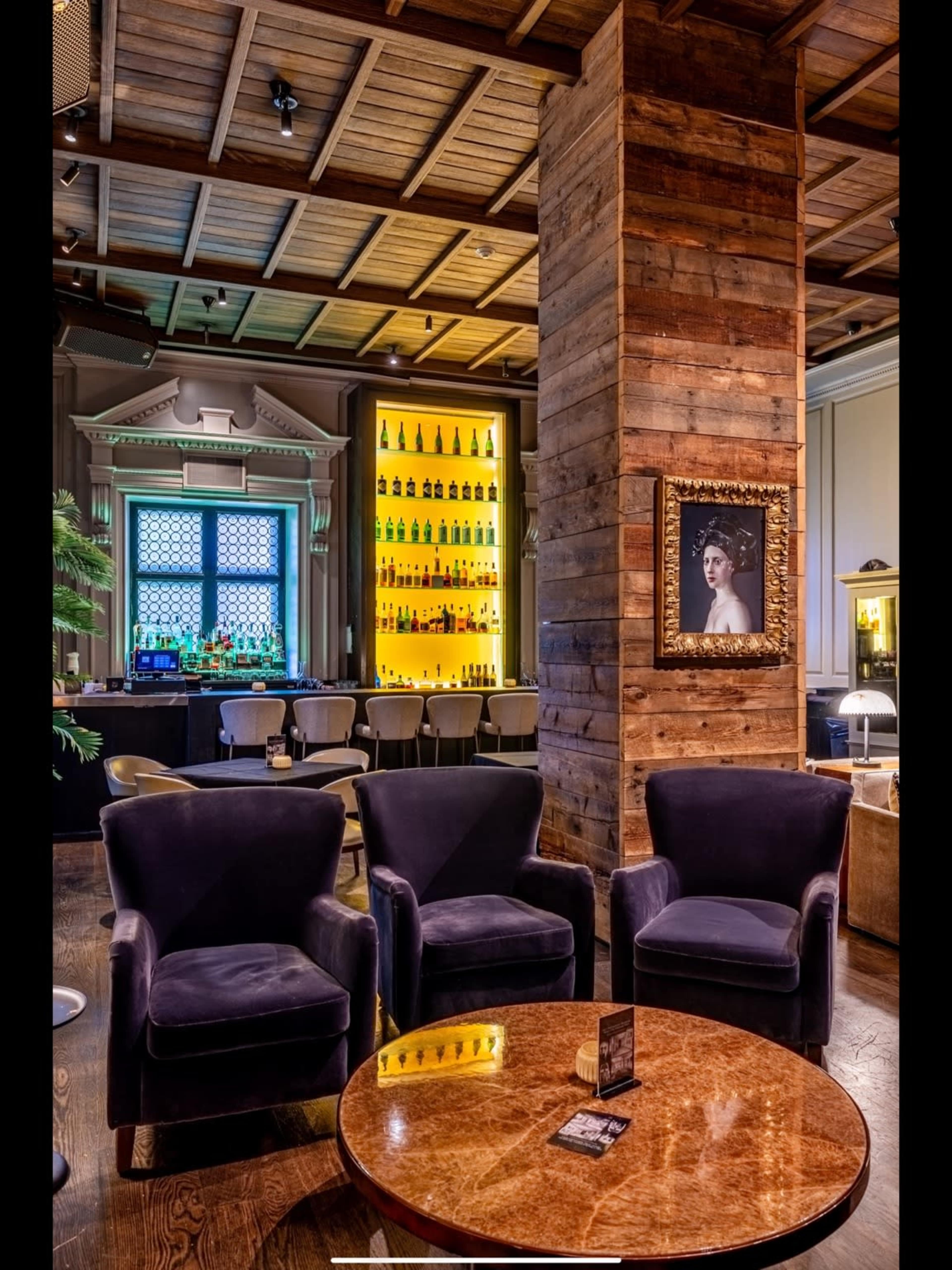 The image shows a cozy seating area in a bar with three plush chairs and a wooden table, beside a well-lit bar featuring a variety of bottles.