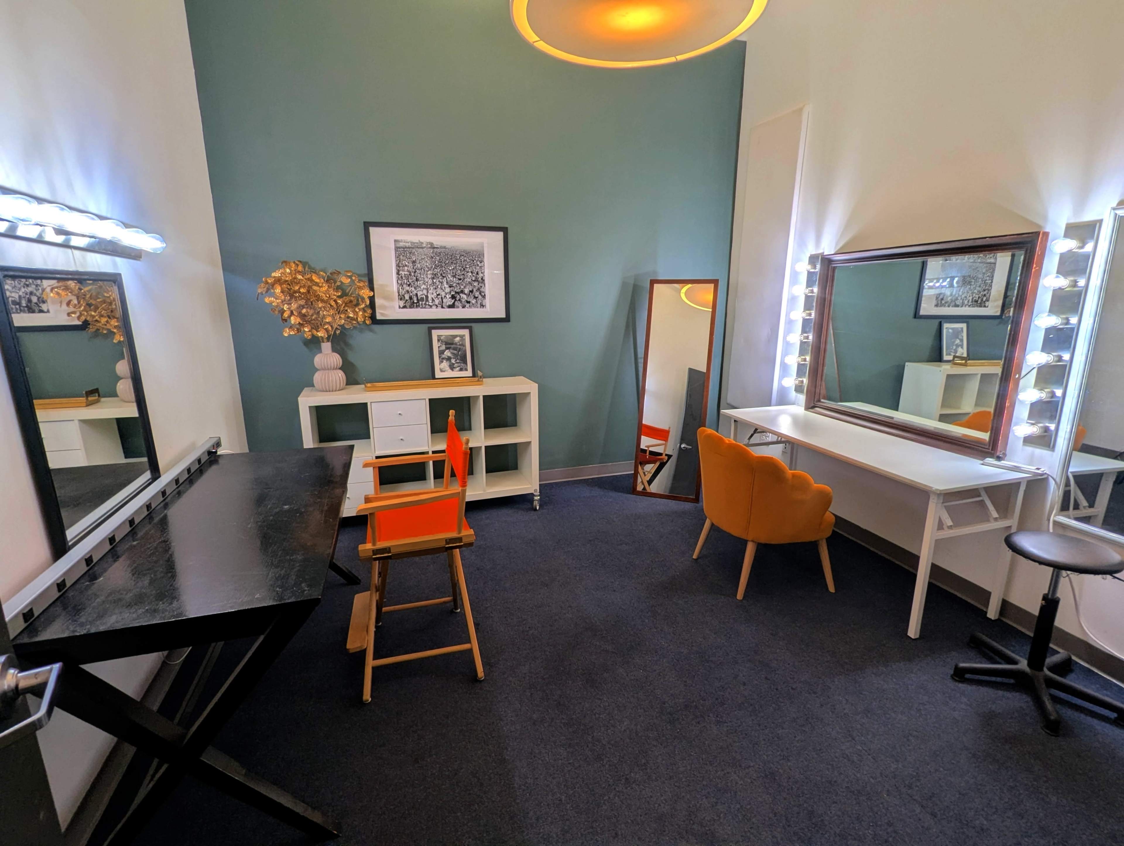 The image shows a well-lit dressing room featuring two mirrors with bulbs, a black table, a wooden chair, and a white desk against a green wall.