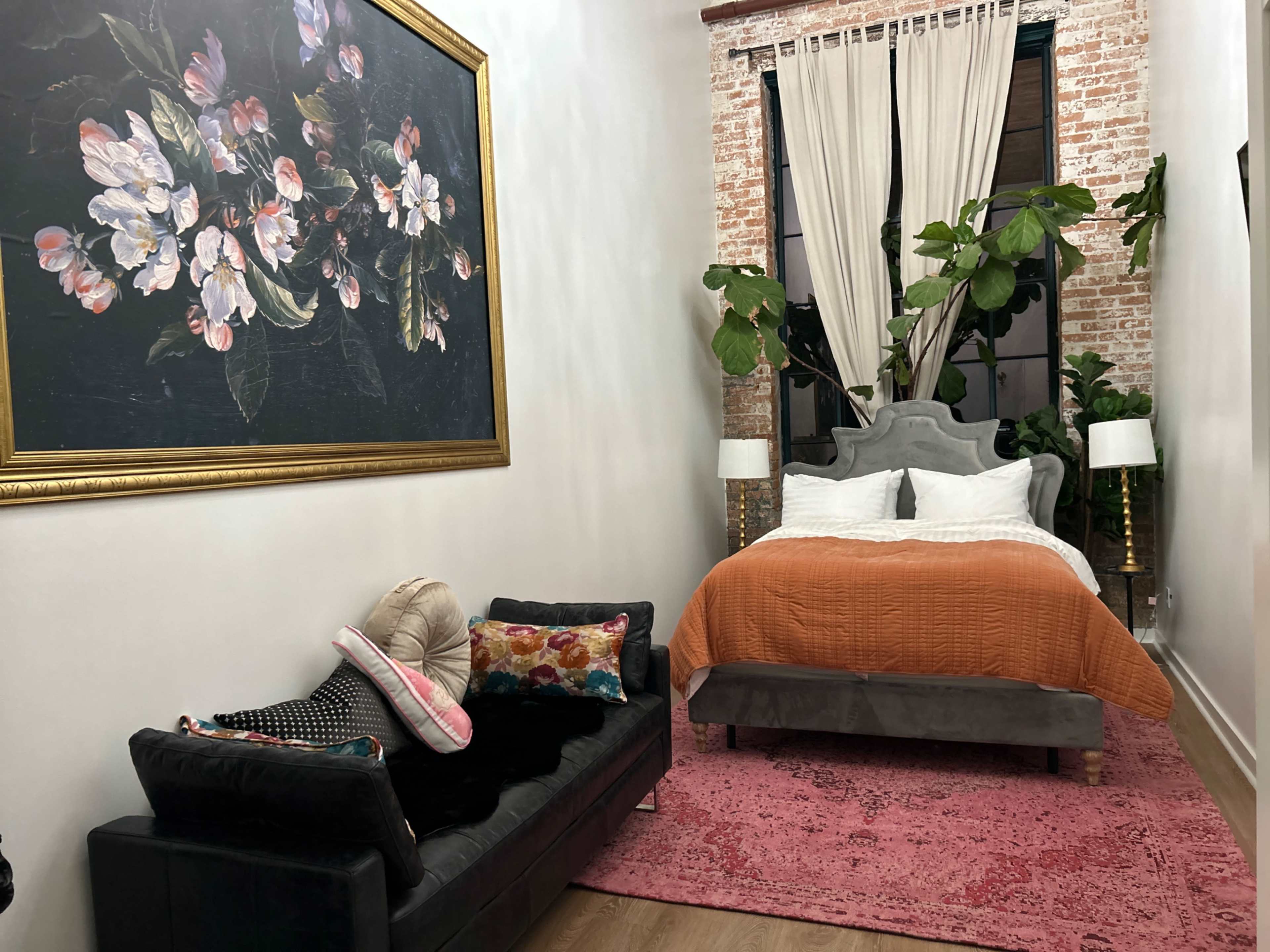 The room features a bed with a rust-colored duvet, adorned with colorful pillows, a large floral painting on the wall, and a green plant near a window.