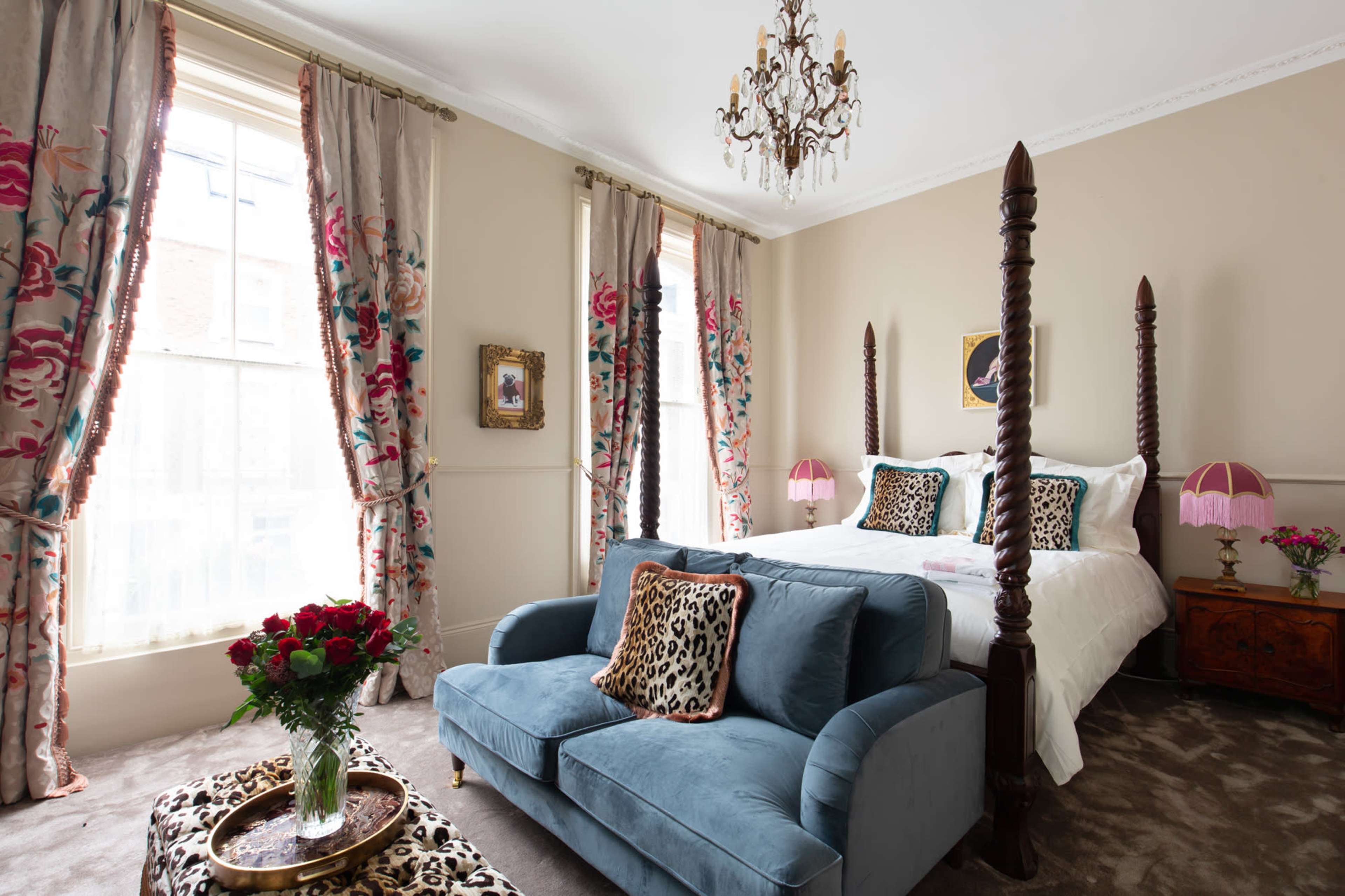 The room features a four-poster bed, a blue sofa, and floral curtains, with a chandelier overhead and a vase of roses on a coffee table.