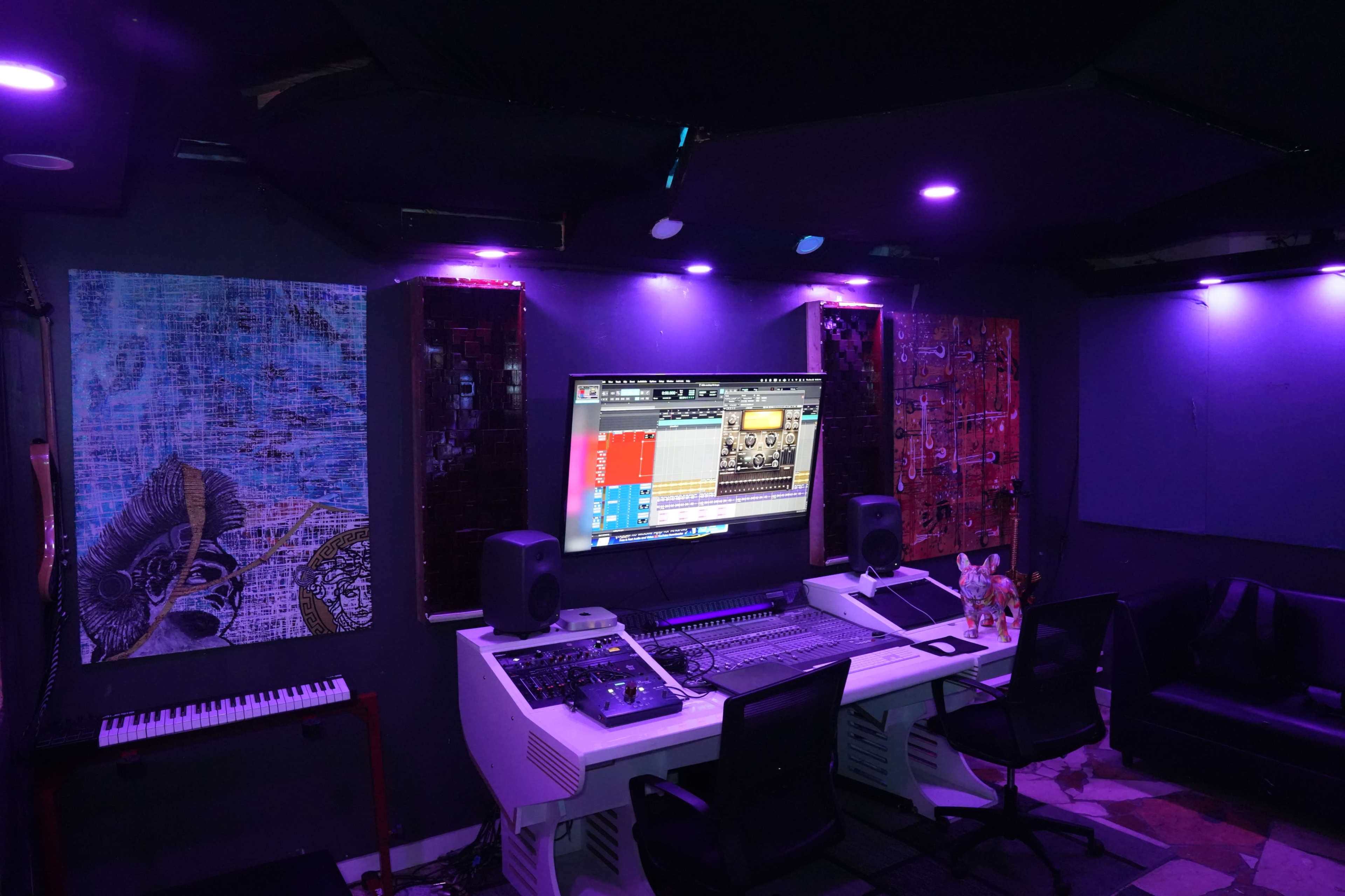 A music production studio features a mixing console, an expansive monitor, and colorful artwork on the walls.