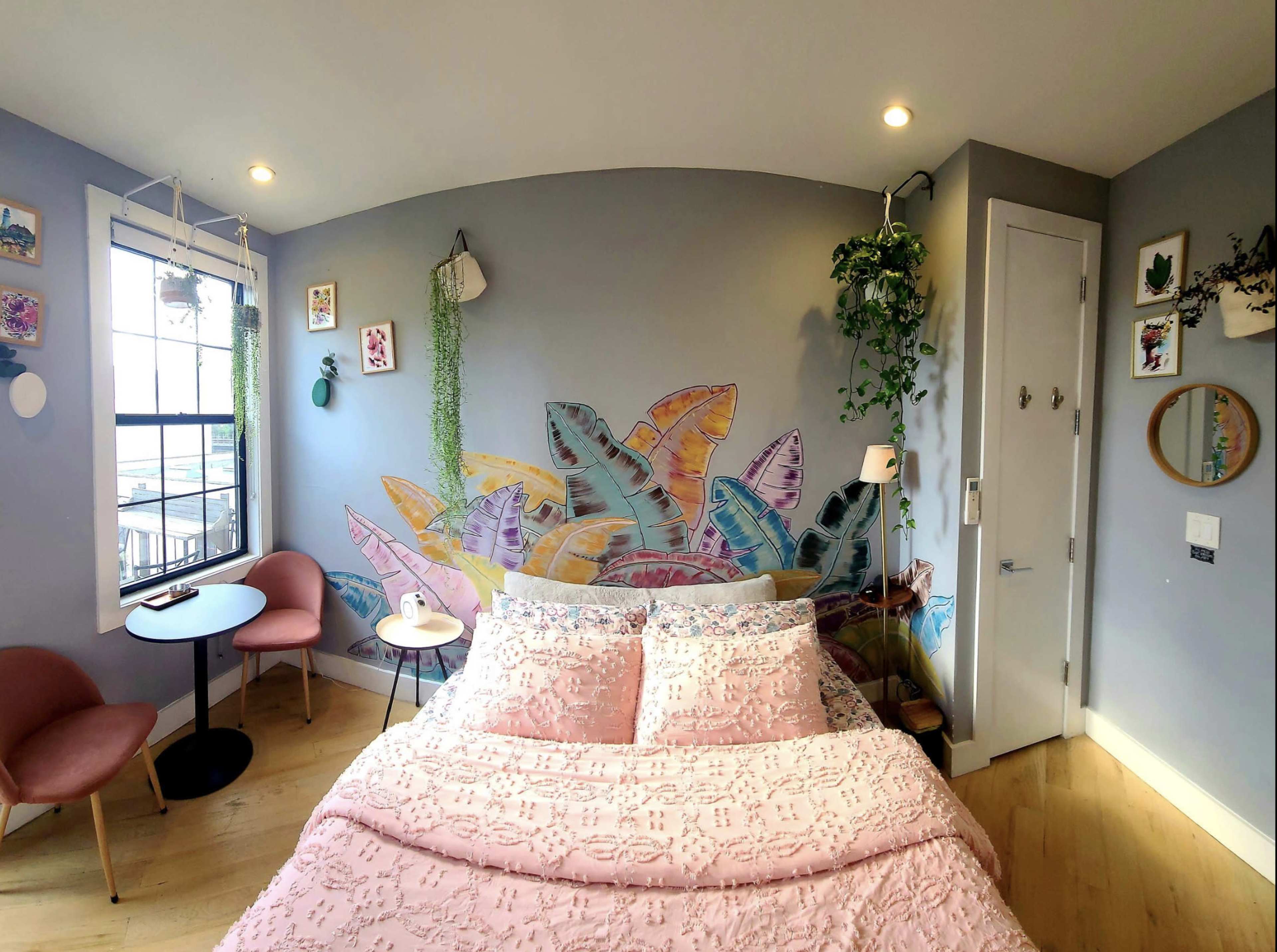 The image shows a cozy bedroom featuring a pastel-colored mural of fish on the wall, a pink bedspread, and two chairs with a small table nearby.