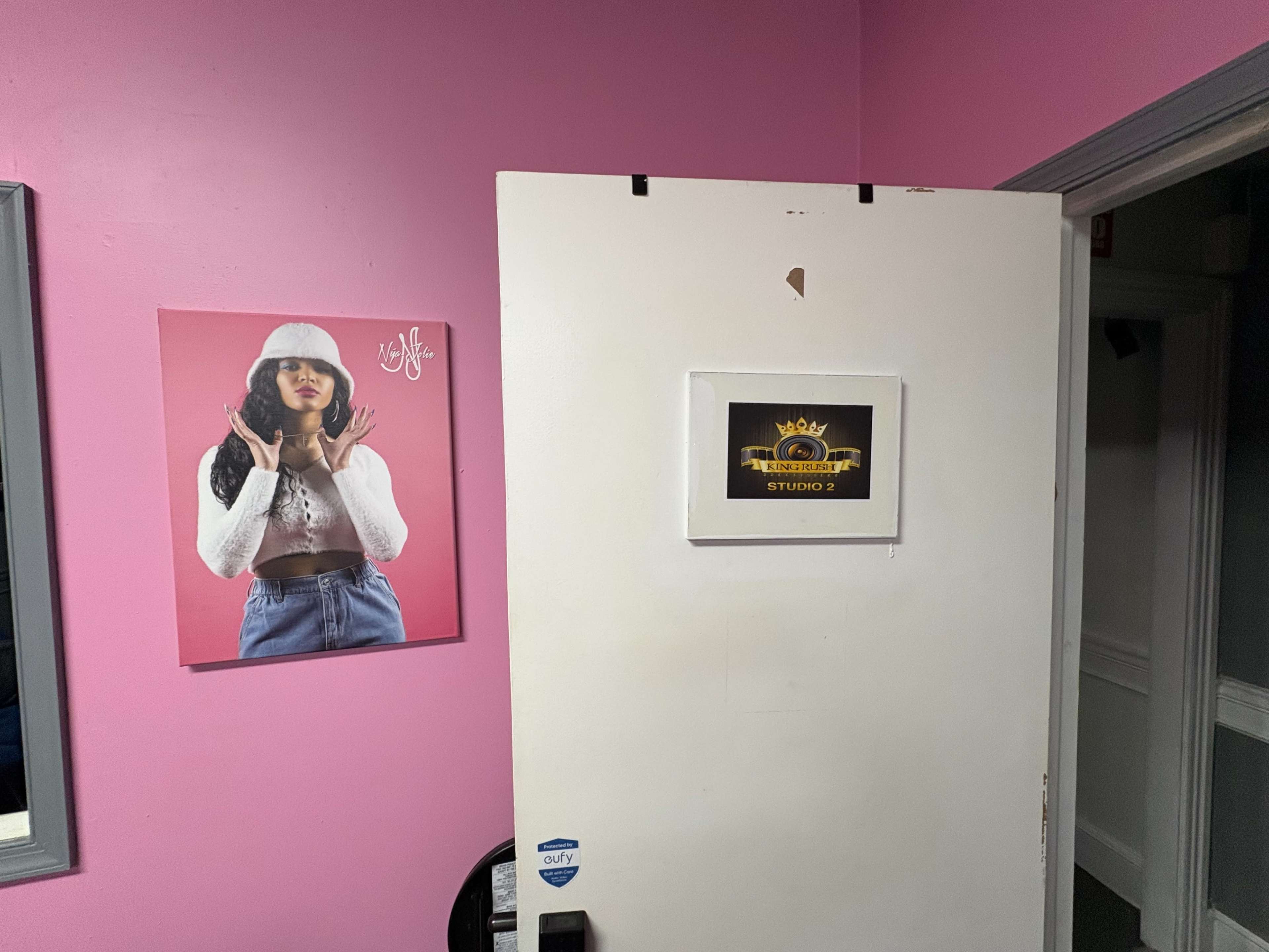 A white door with a sign labeled "STUDIO 2" is partially open in a pink room, featuring a framed portrait of a woman wearing a white hat.