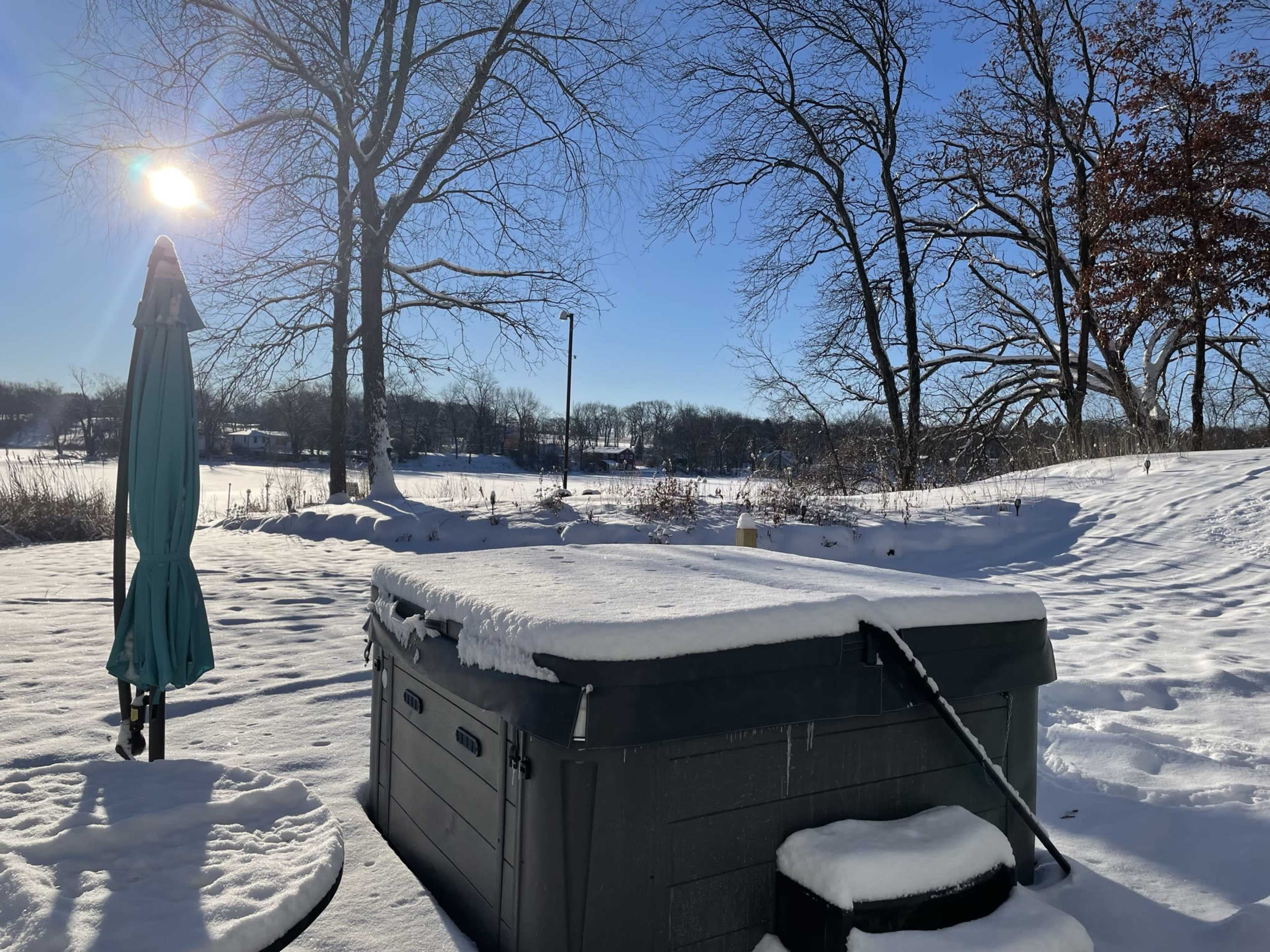A hot tub covered in snow sits in a backyard with bare trees and a clear blue sky.