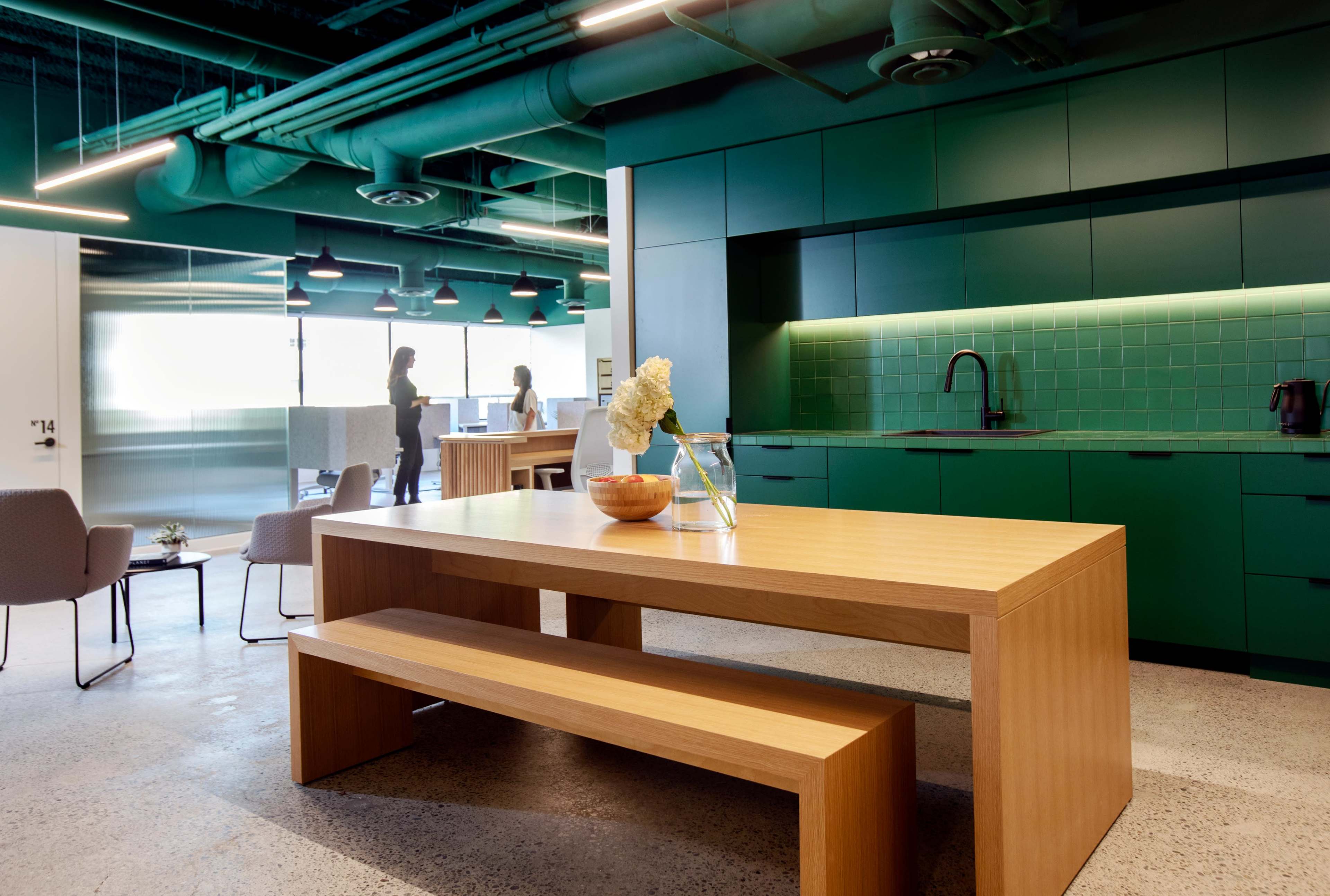 A modern office kitchen area featuring a wooden table and bench, green cabinetry, and two people engaged in conversation in the background.