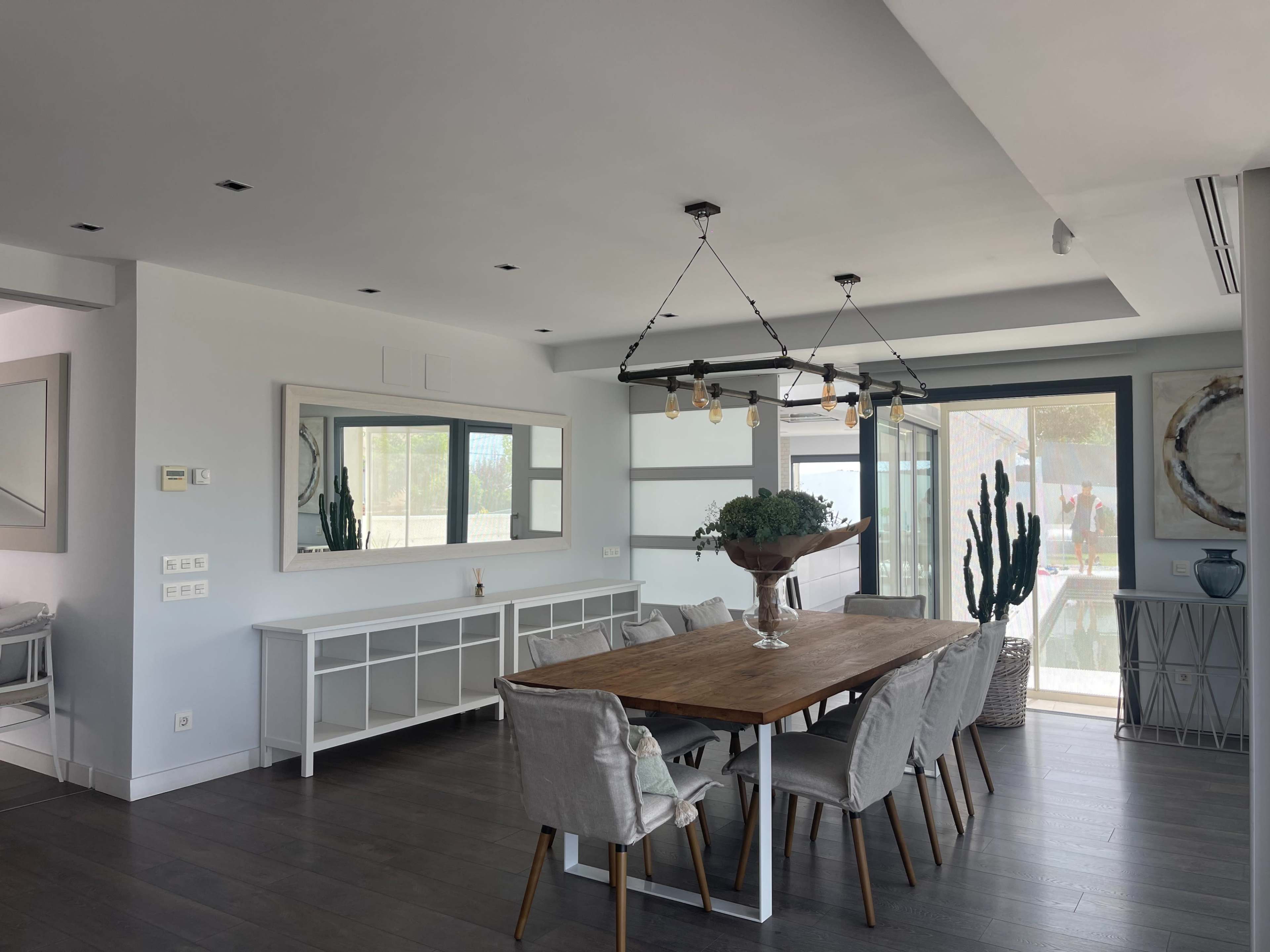 A modern dining area featuring a wooden table surrounded by upholstered chairs, with large windows and plants enhancing the space.