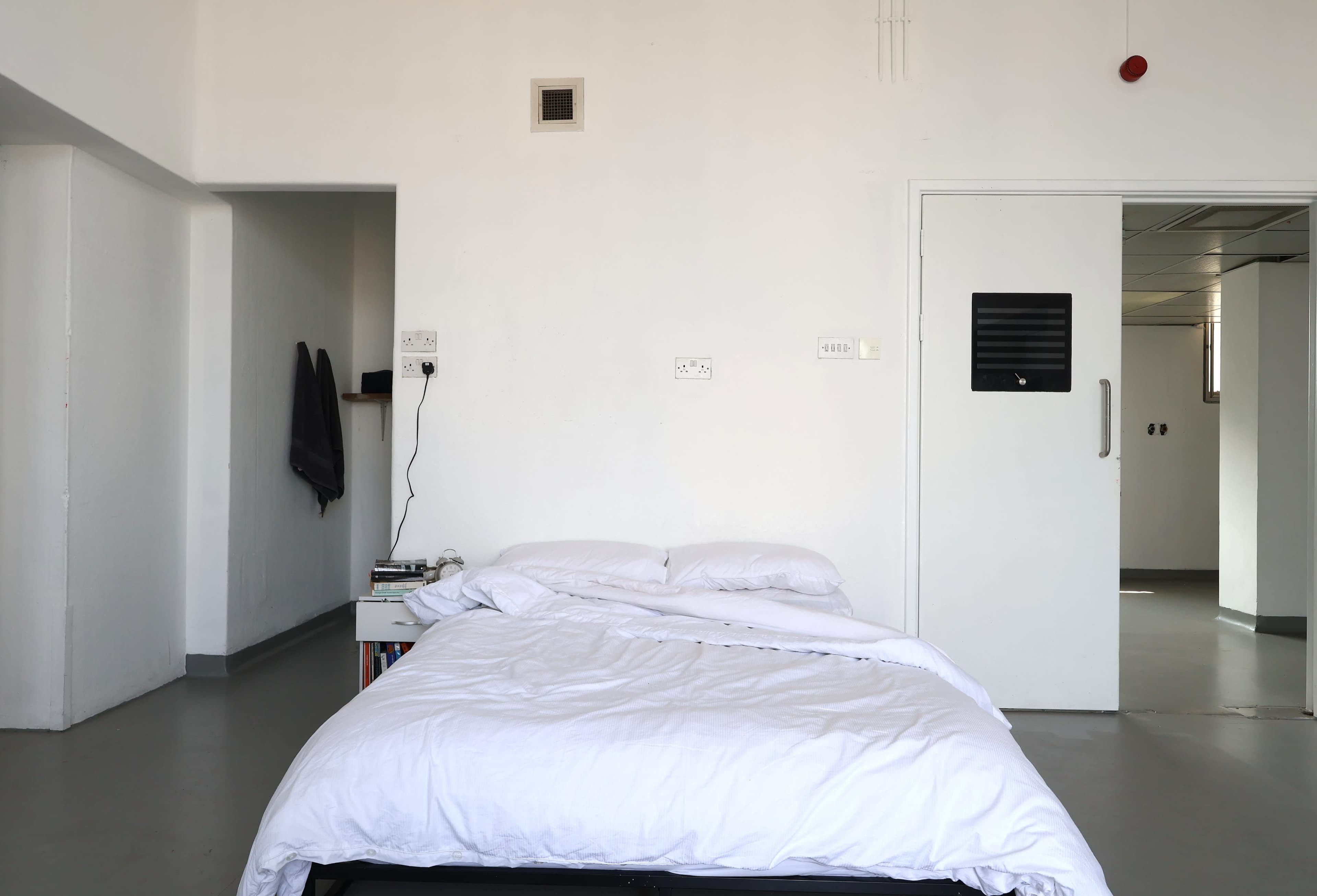 A bed with a white duvet is positioned in a sparsely furnished room with plain white walls.