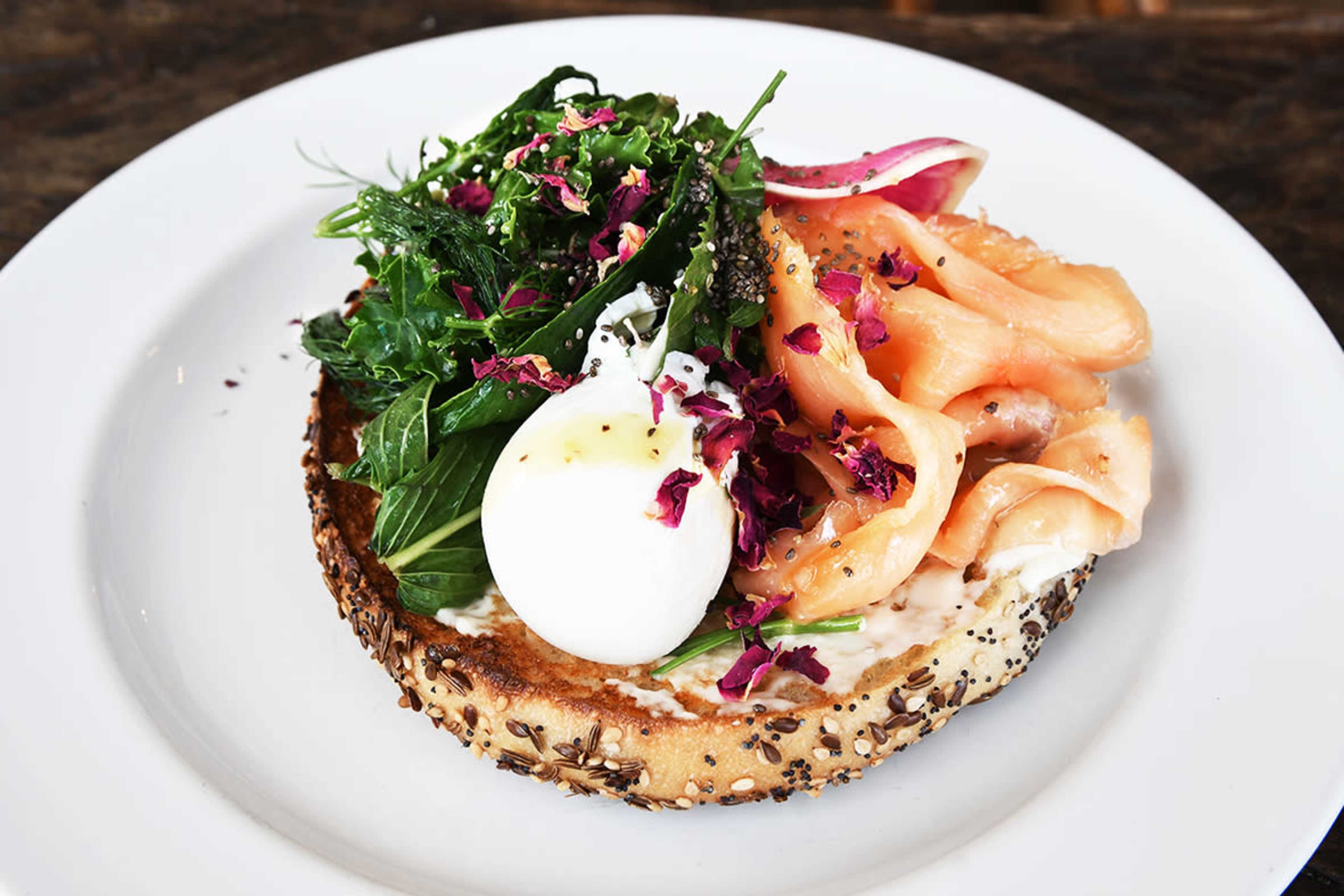 The image shows a toasted bagel topped with smoked salmon, a poached egg, greens, and a garnish of rose petals.