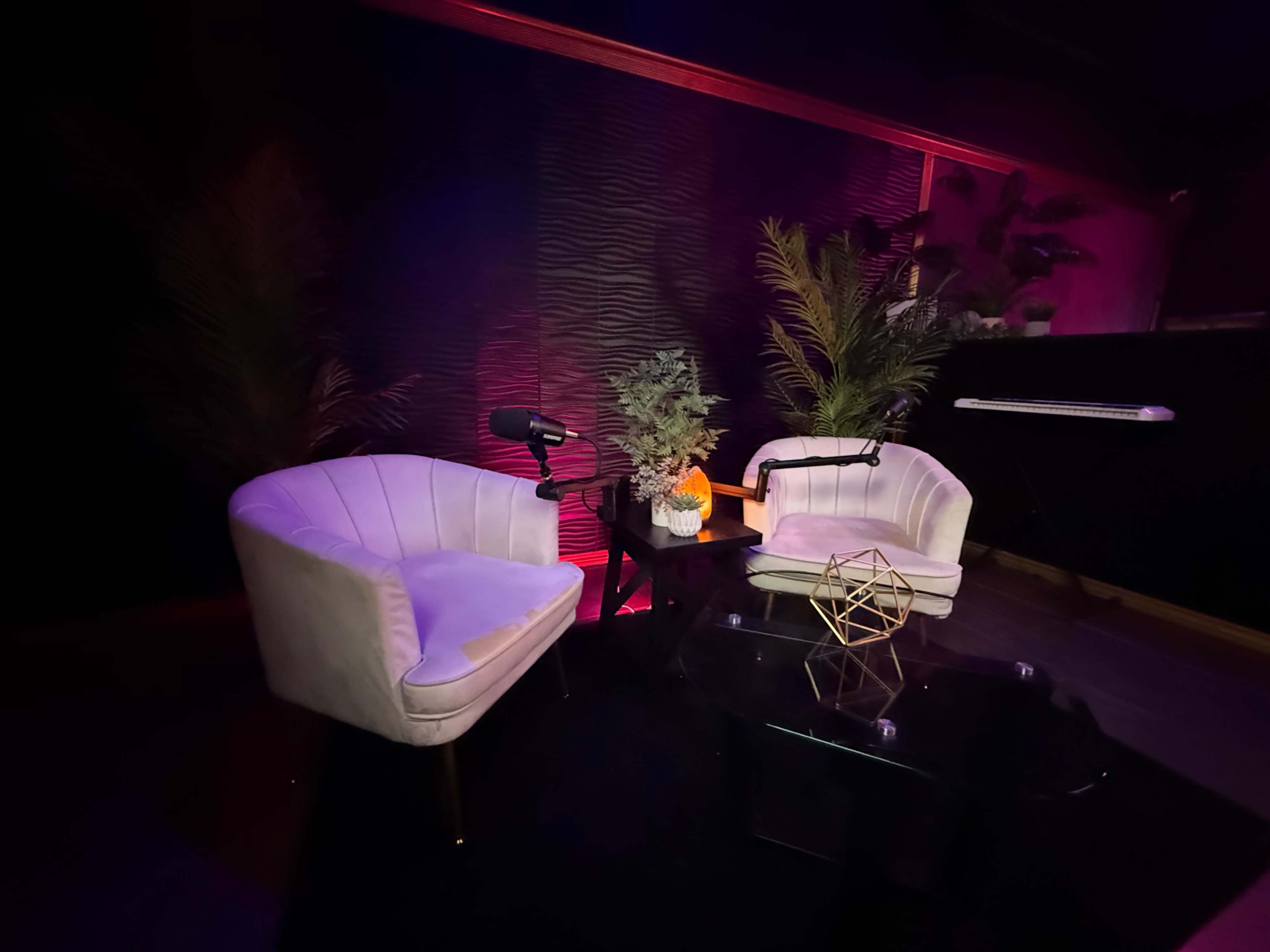 The image shows a small, darkened studio setup featuring two white armchairs facing a glass table with decorative items and plants in the background.