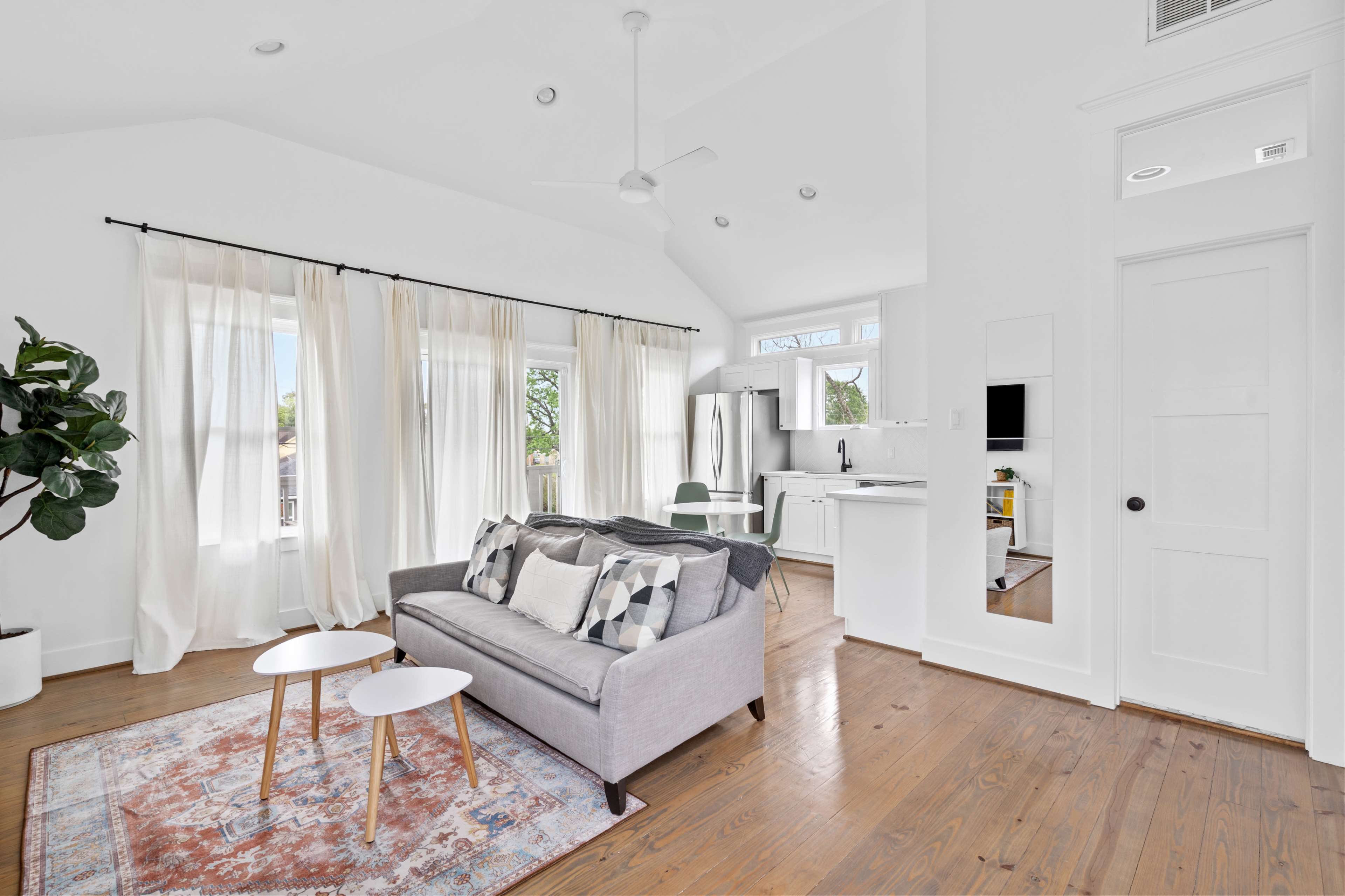 The image shows a bright, modern living room with a gray sofa, two small white tables, and large windows covered with light curtains.