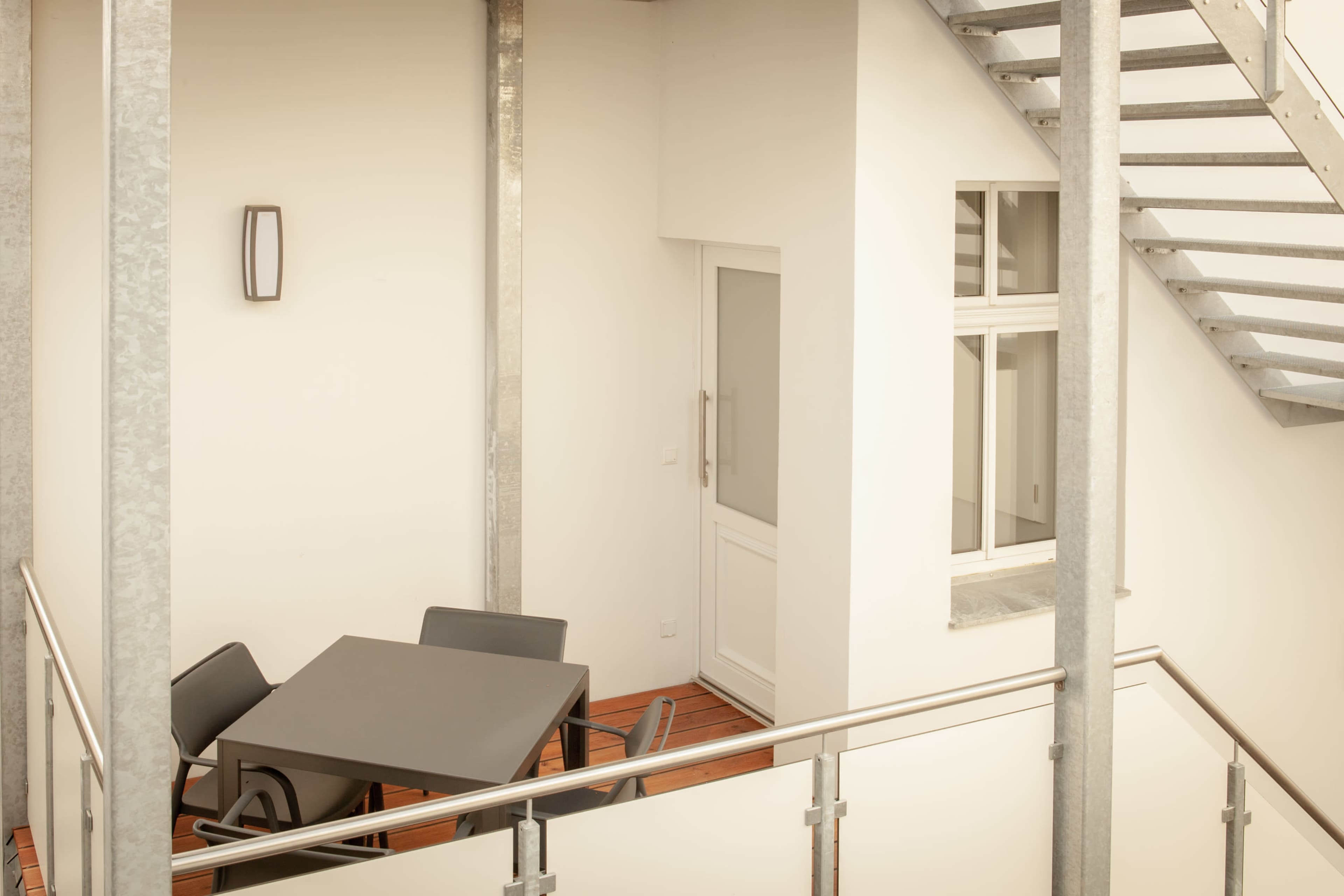 The image shows a modern balcony with a table and chairs, surrounded by metal railings and a staircase leading to an upper level.
