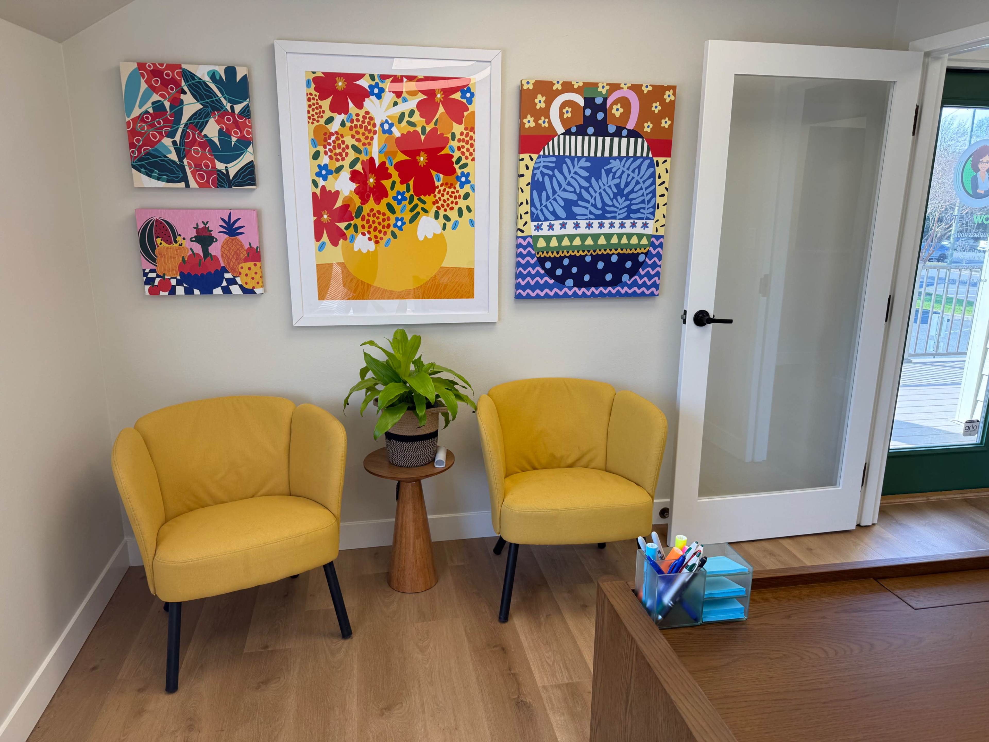 The image shows a bright interior space featuring two yellow armchairs, a wooden side table with a potted plant, and colorful artwork on the walls.