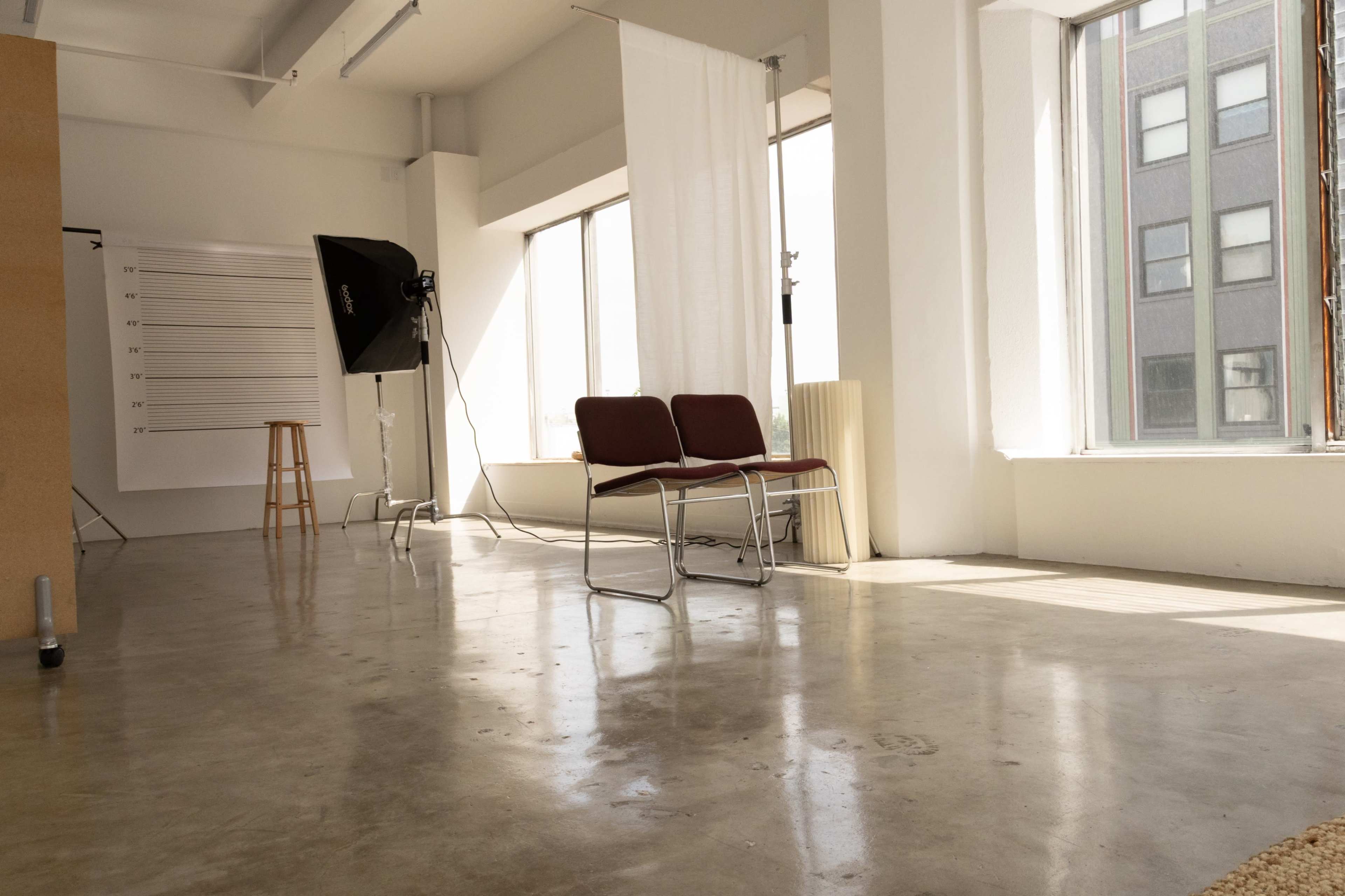 The image shows a spacious, well-lit studio with two red chairs, a photography light setup, and large windows letting in natural light.