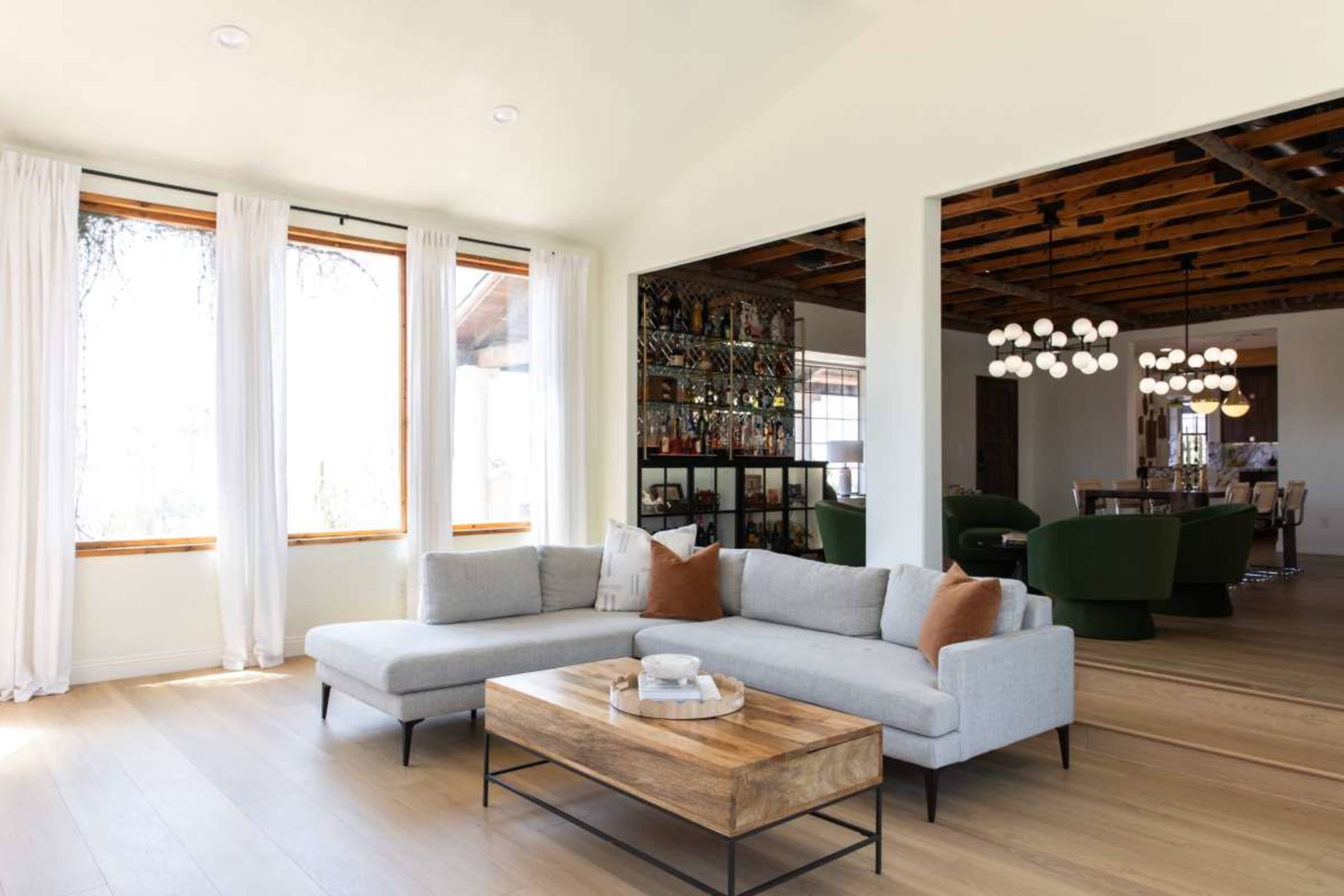 The image shows a modern living room with a light gray sectional sofa, a wooden coffee table, and large windows allowing natural light to flow in.
