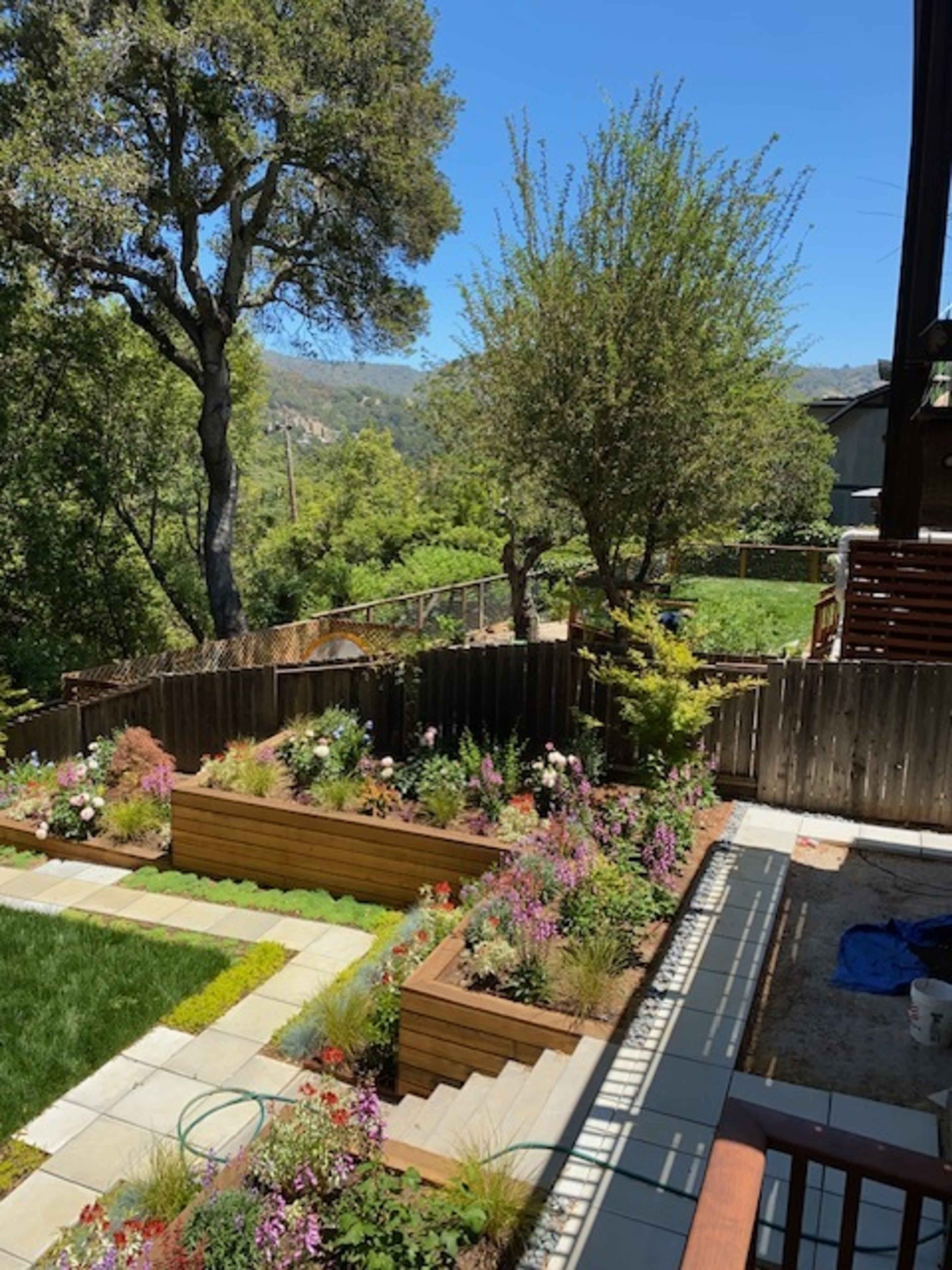 A landscaped backyard with raised garden beds filled with various plants and flowers, surrounded by trees and a scenic hill in the background.