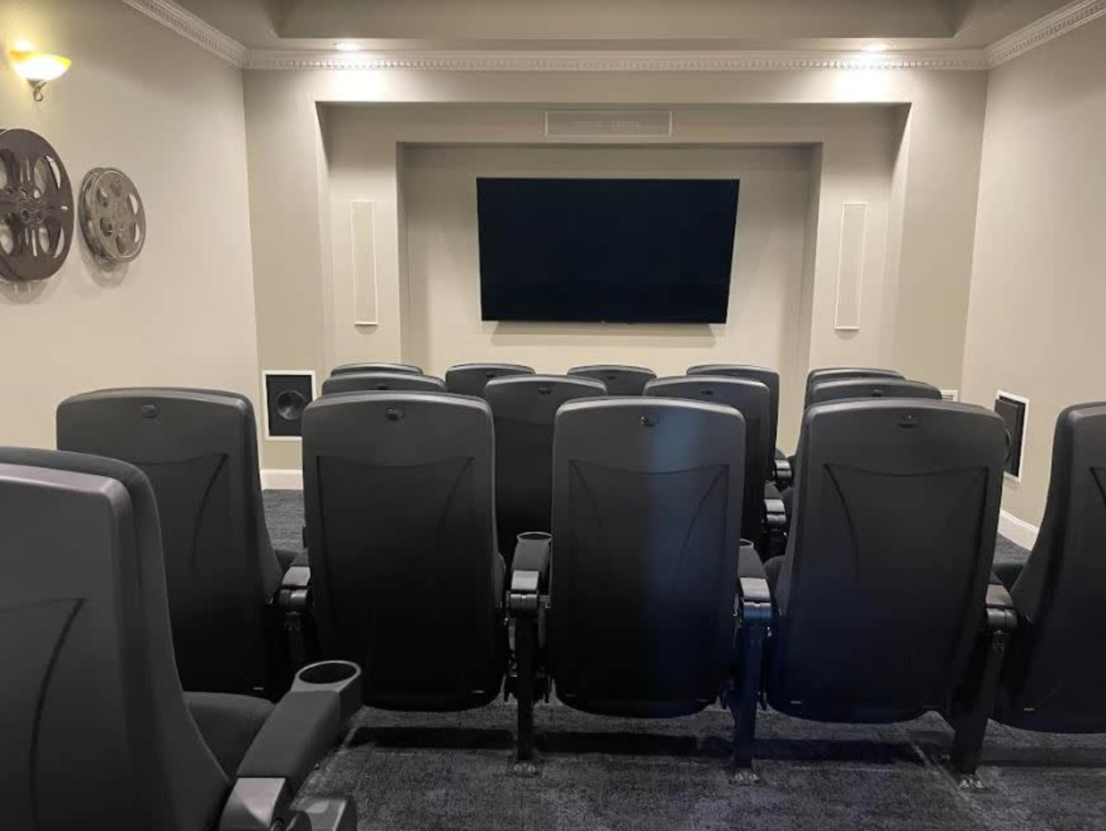 A home theater room with black reclining chairs arranged in rows facing a large flat-screen TV mounted on the wall.