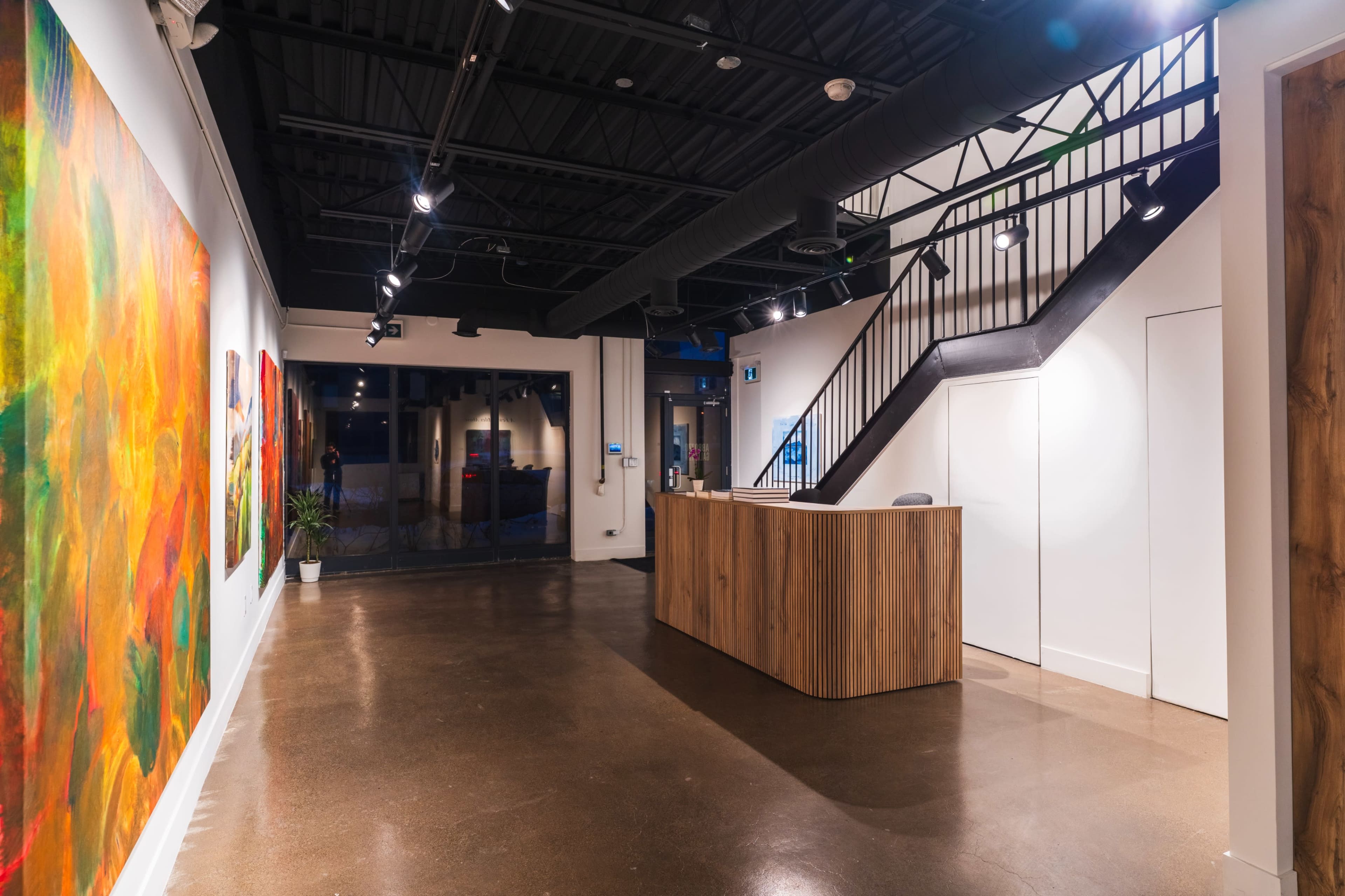 The image shows a modern reception area with an art-filled wall, wooden reception desk, and a staircase leading to an upper level.