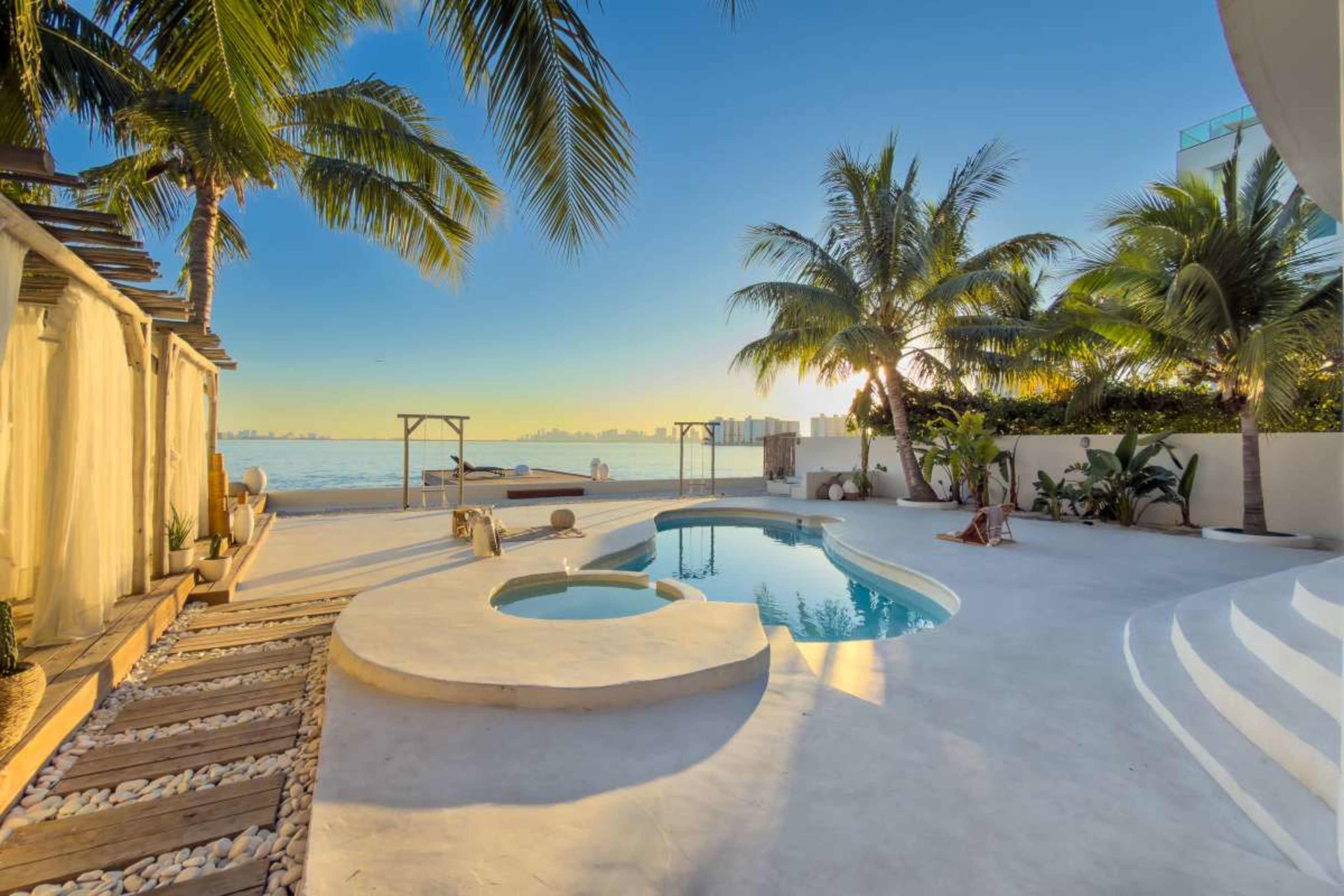 A swimming pool with a circular hot tub is surrounded by palm trees and a deck overlooking the calm water in the distance.