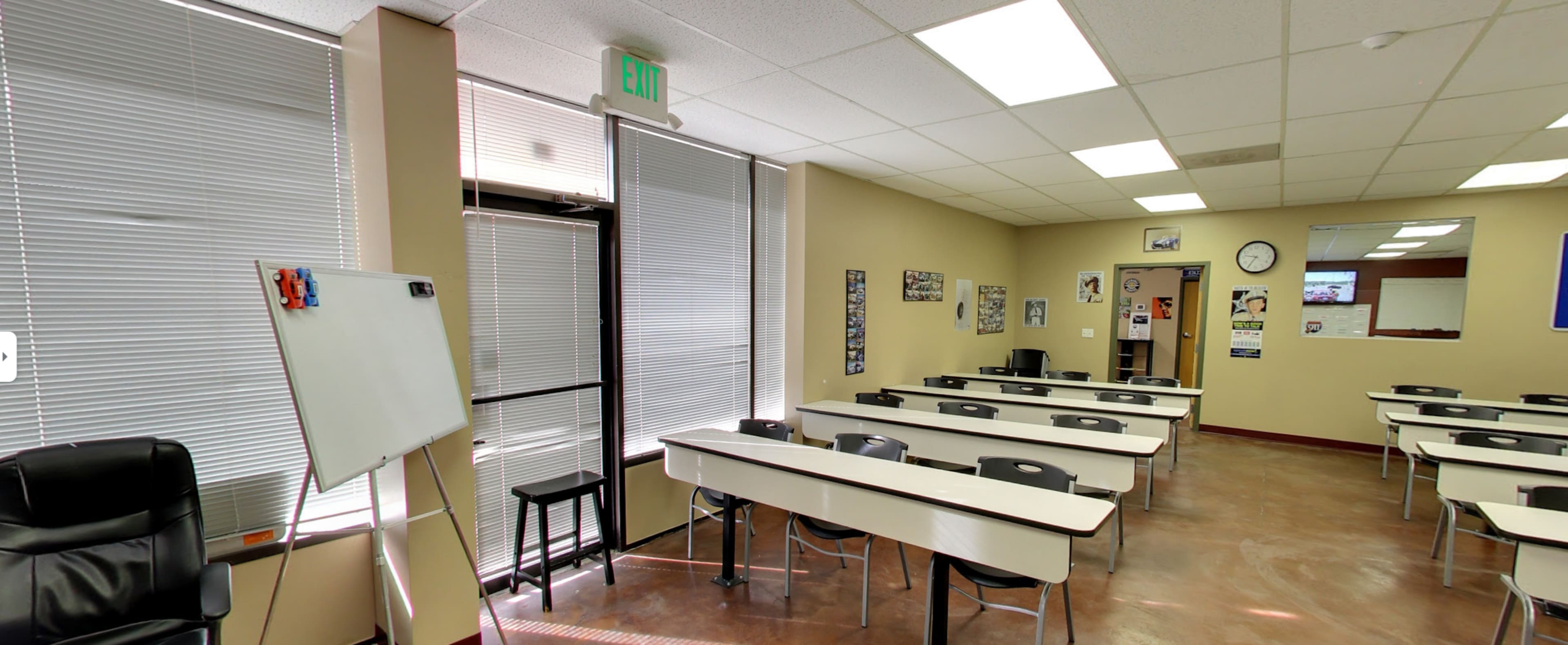 The image shows a classroom with rows of desks, a whiteboard, and large windows allowing light to enter.
