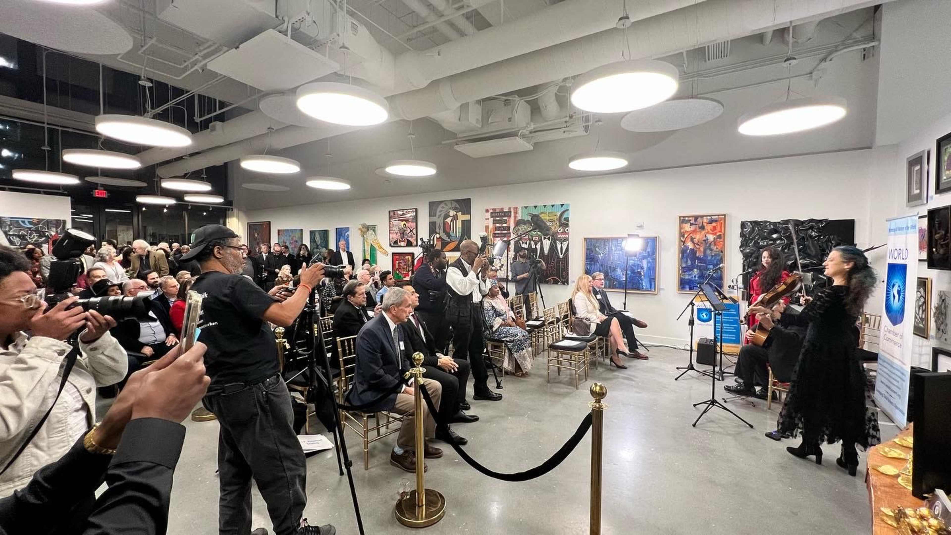 A diverse audience watches a live musical performance while some attendees capture the moment on their cameras in an art gallery setting.