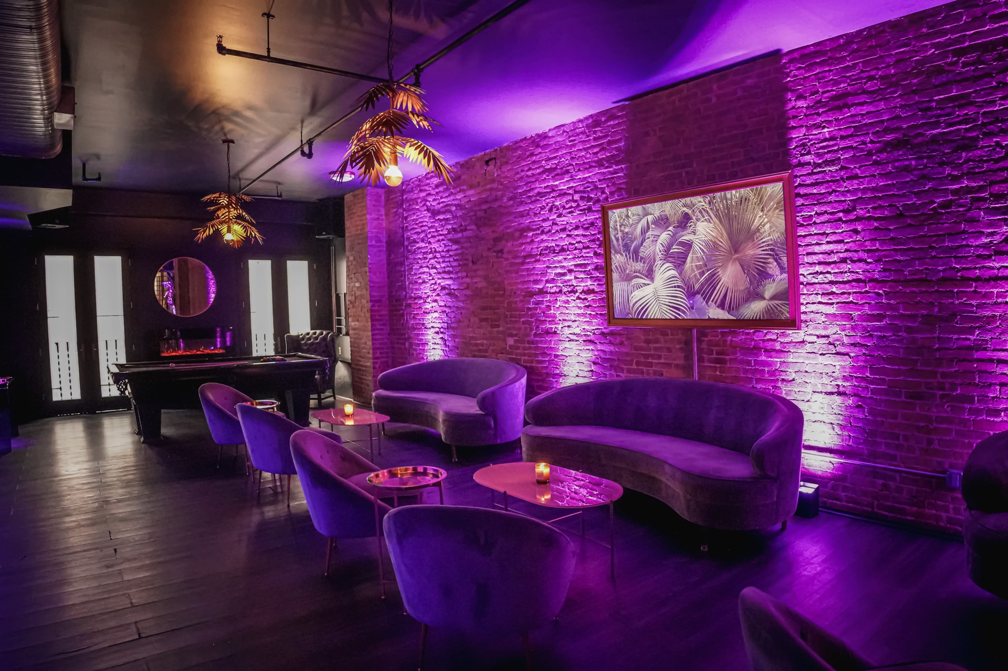 The image shows a dimly lit bar with purple lighting, featuring curved sofas, small tables, and a framed picture of palm leaves on the brick wall.