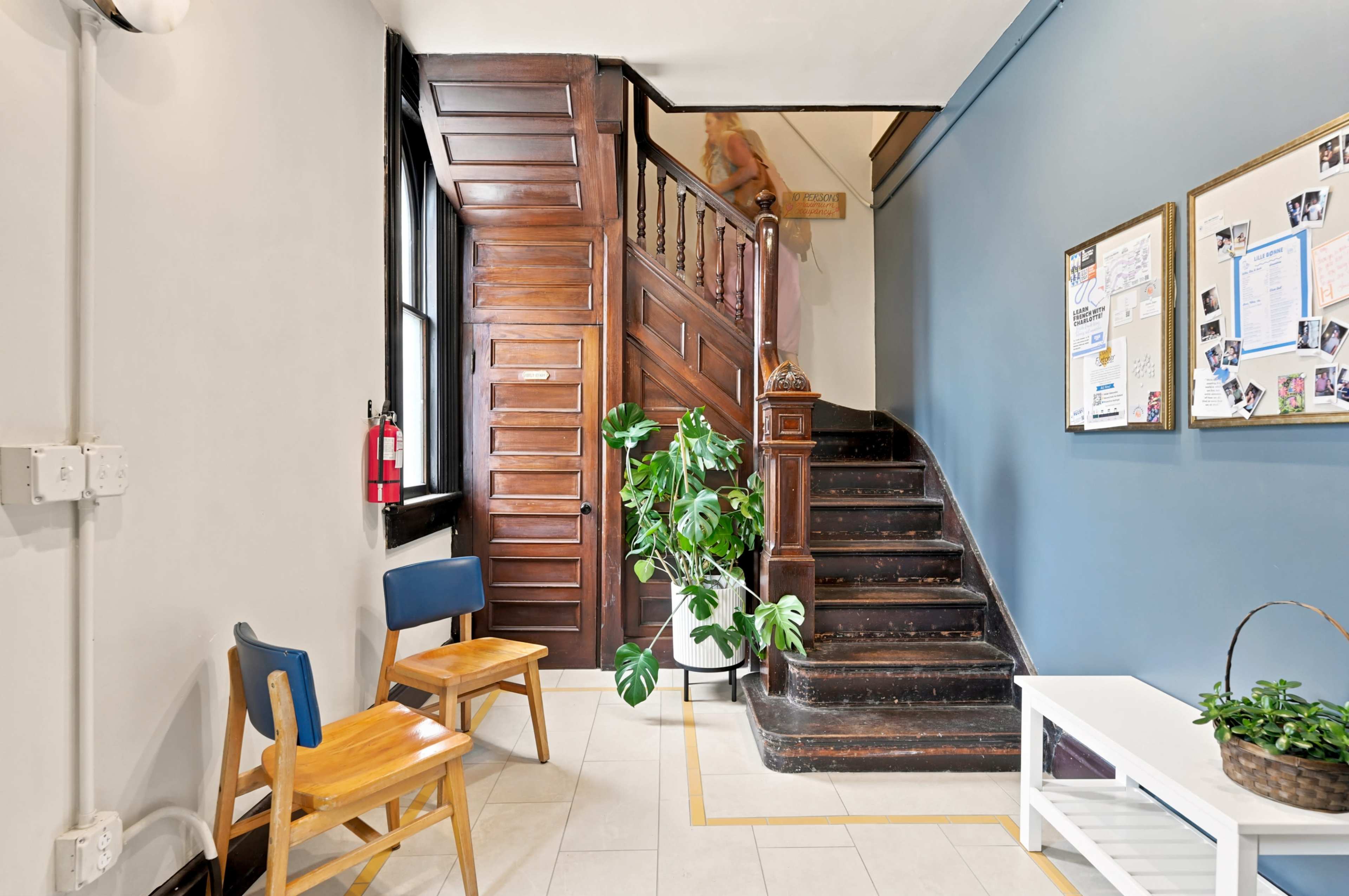 The image shows an interior hallway featuring a wooden staircase, a potted plant, and two wooden chairs beside a white coffee table.