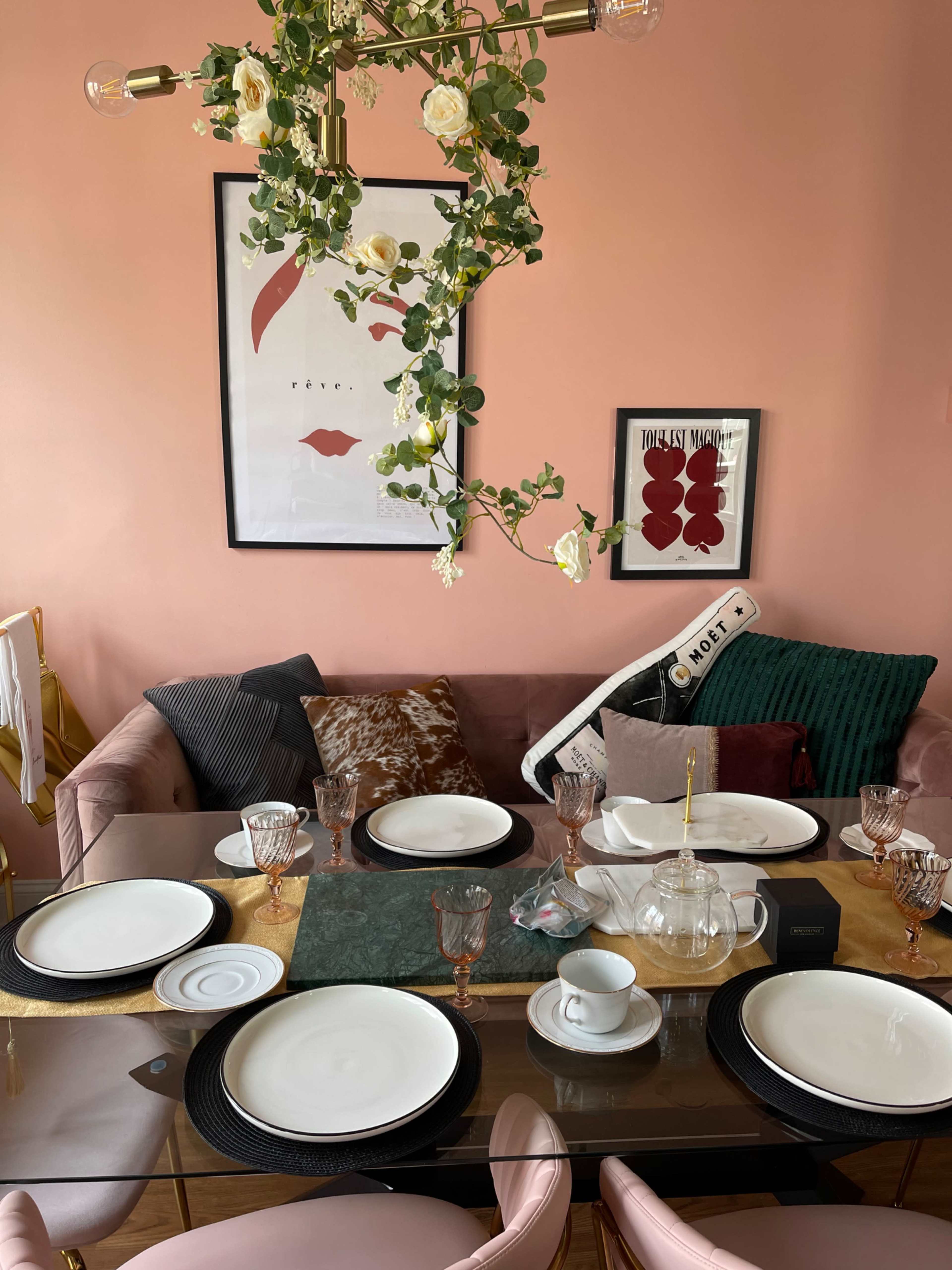 A dining table is set with white plates and glassware, surrounded by a pink wall and a couch adorned with cushions.