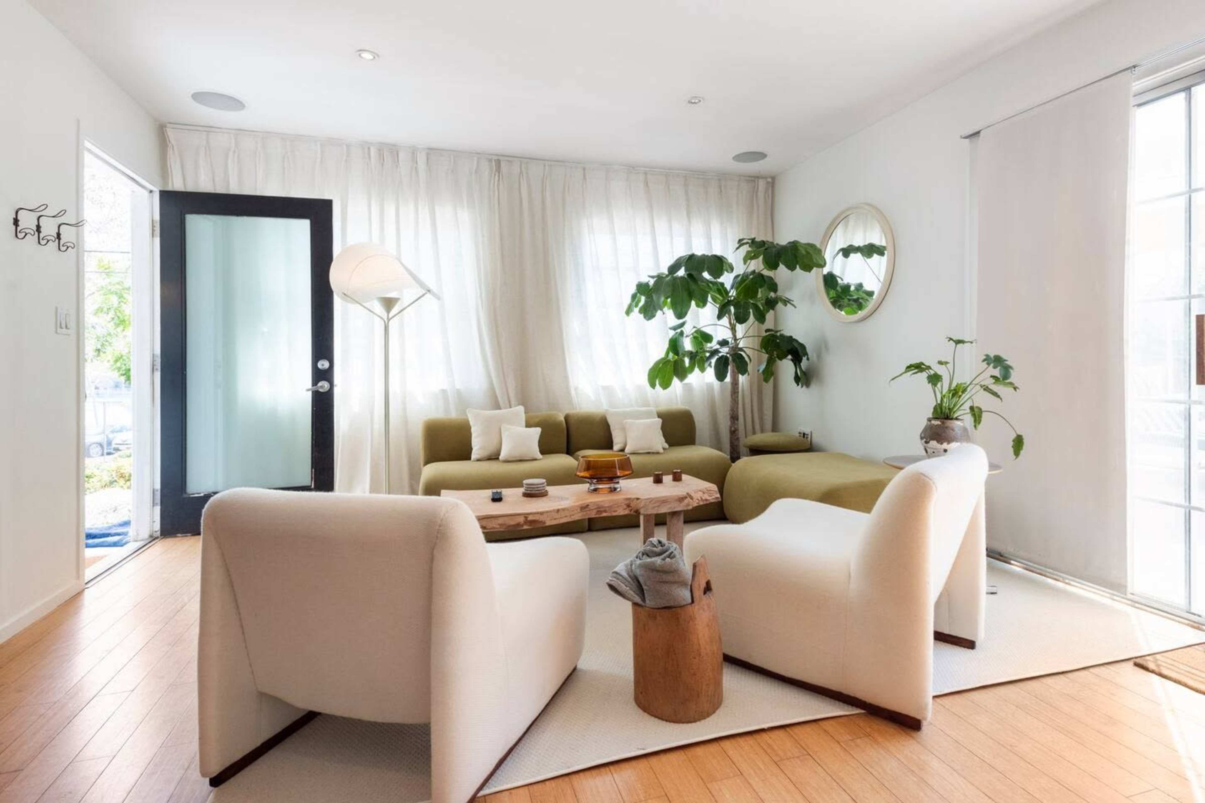 A modern living room with a light-colored sofa, two white chairs, a wooden coffee table, and large windows covered with sheer curtains.