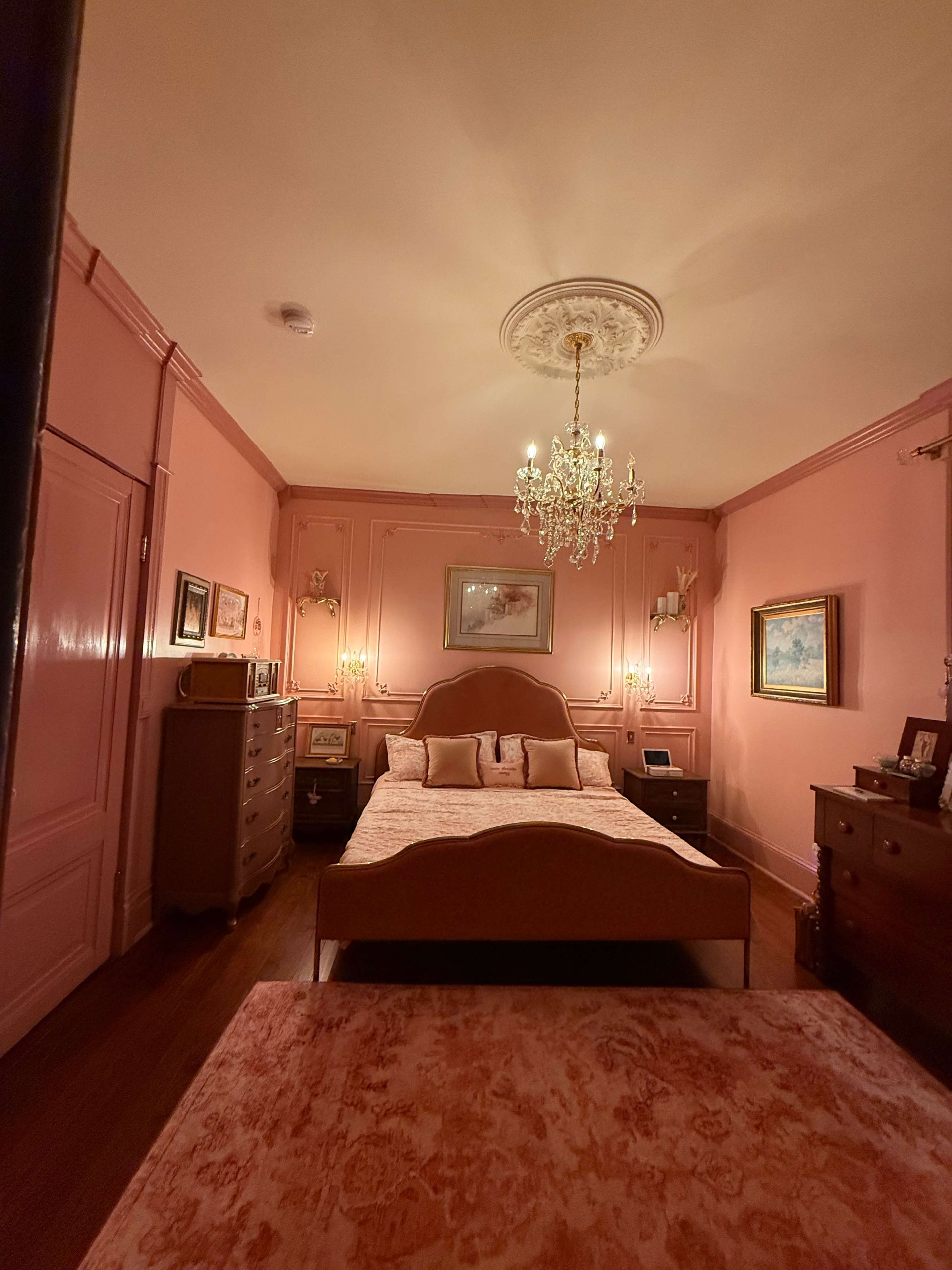 The image shows a softly lit bedroom with pink walls, a large bed at the center, and vintage furniture arranged around the room.