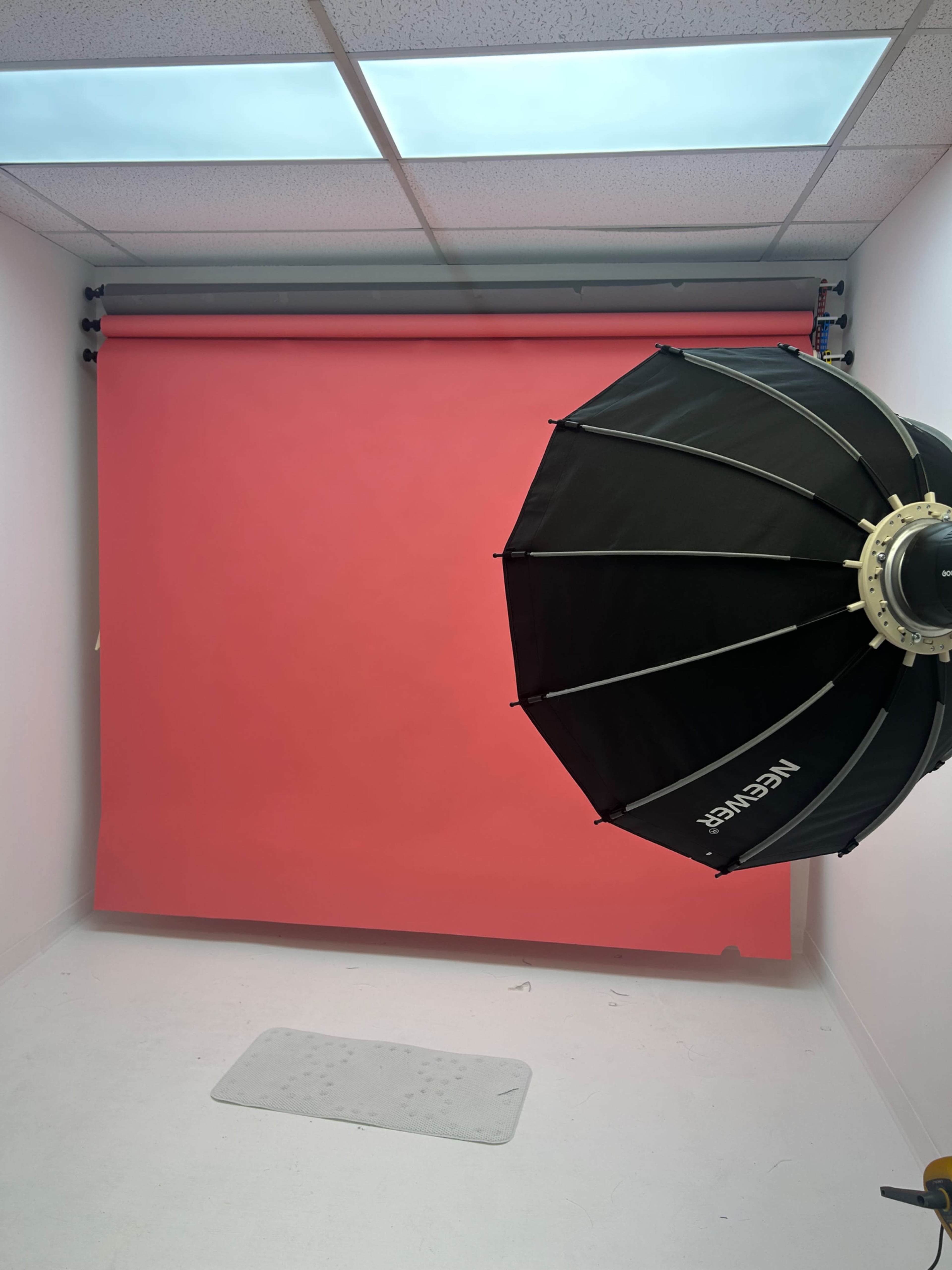 The image shows a photography studio setup featuring a pink backdrop and a black softbox light in the corner.