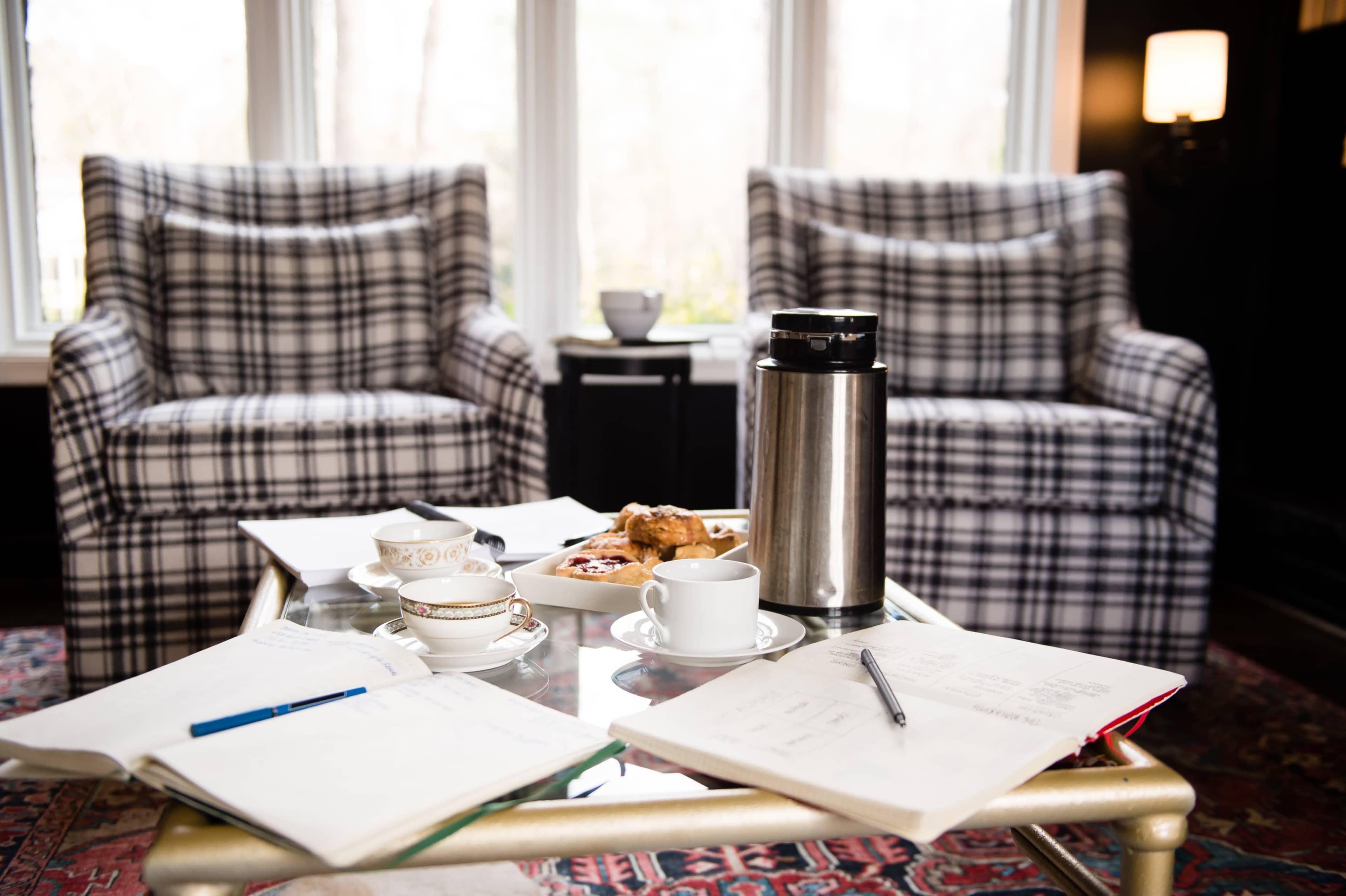 A cozy seating area features two plaid armchairs, a coffee thermos, pastries on a plate, and scattered notebooks on a glass coffee table.