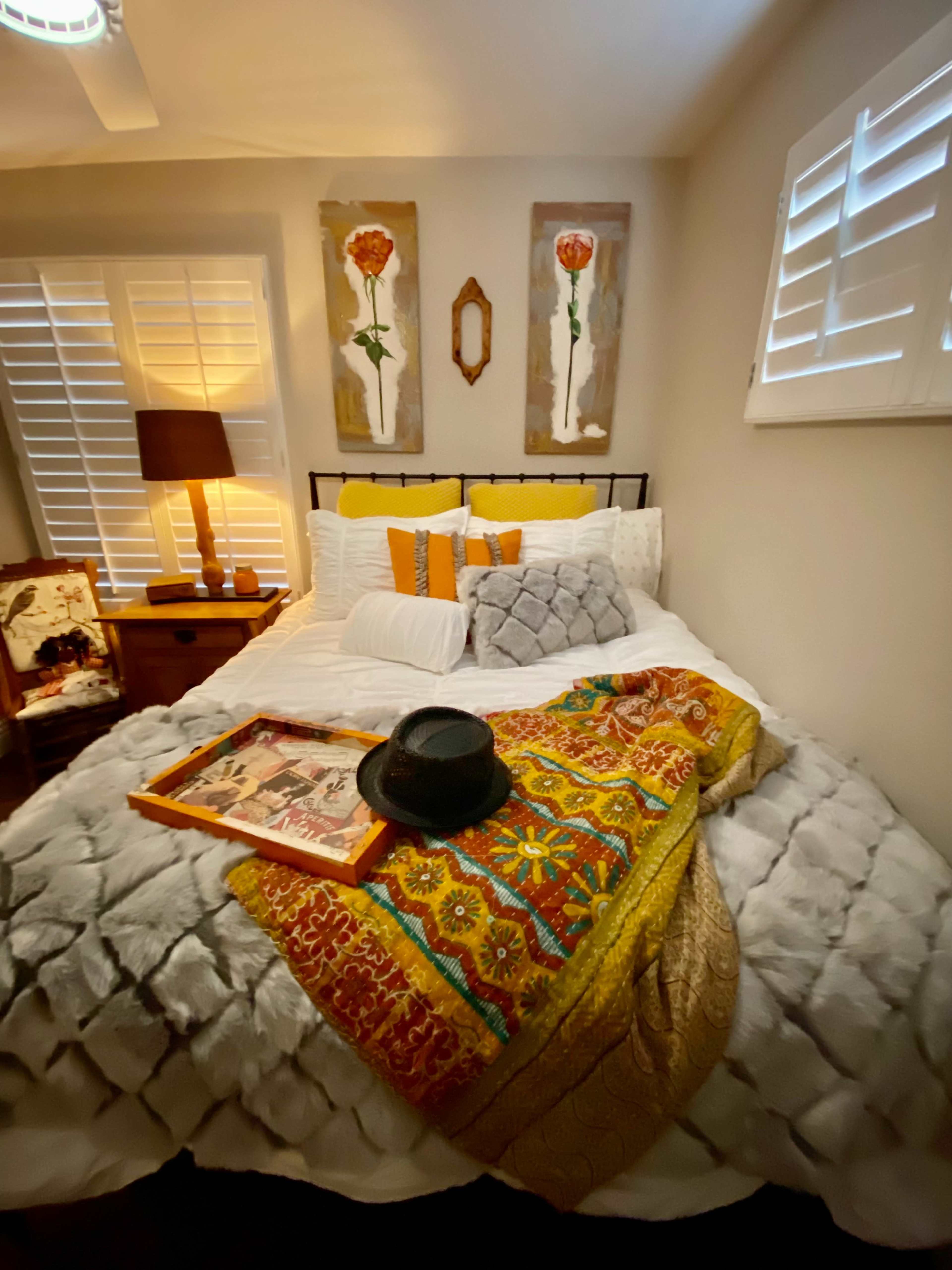A neatly arranged bed with colorful pillows and a throw blanket is positioned in a bright room decorated with wall art and a bedside lamp.