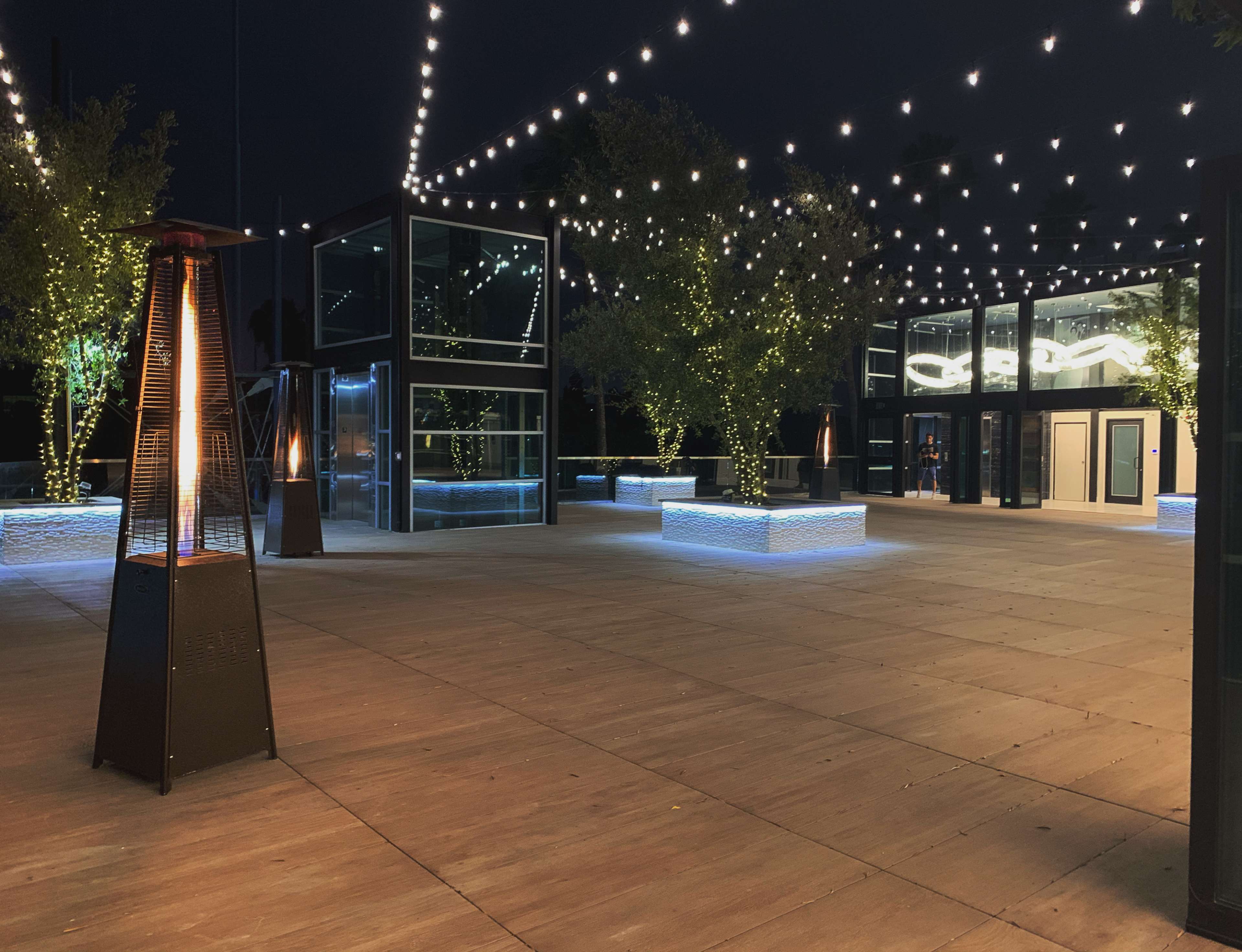 An outdoor venue at night, featuring a wooden floor, illuminated trees, string lights overhead, and modern glass structures.